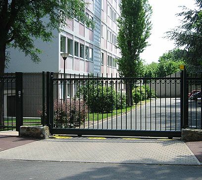 Portail métallique noir fermé devant un immeuble d'appartements gris et une allée