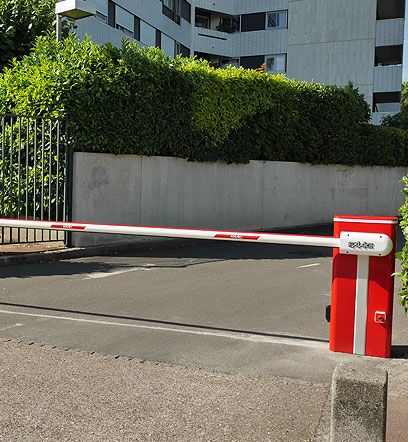 Barrière de parking rouge et blanche bloquant une allée devant une haie et un bâtiment