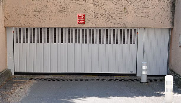 Porte de garage blanche fermée sur un bâtiment beige, avec un panneau rouge au-dessus et une petite borne à proximité