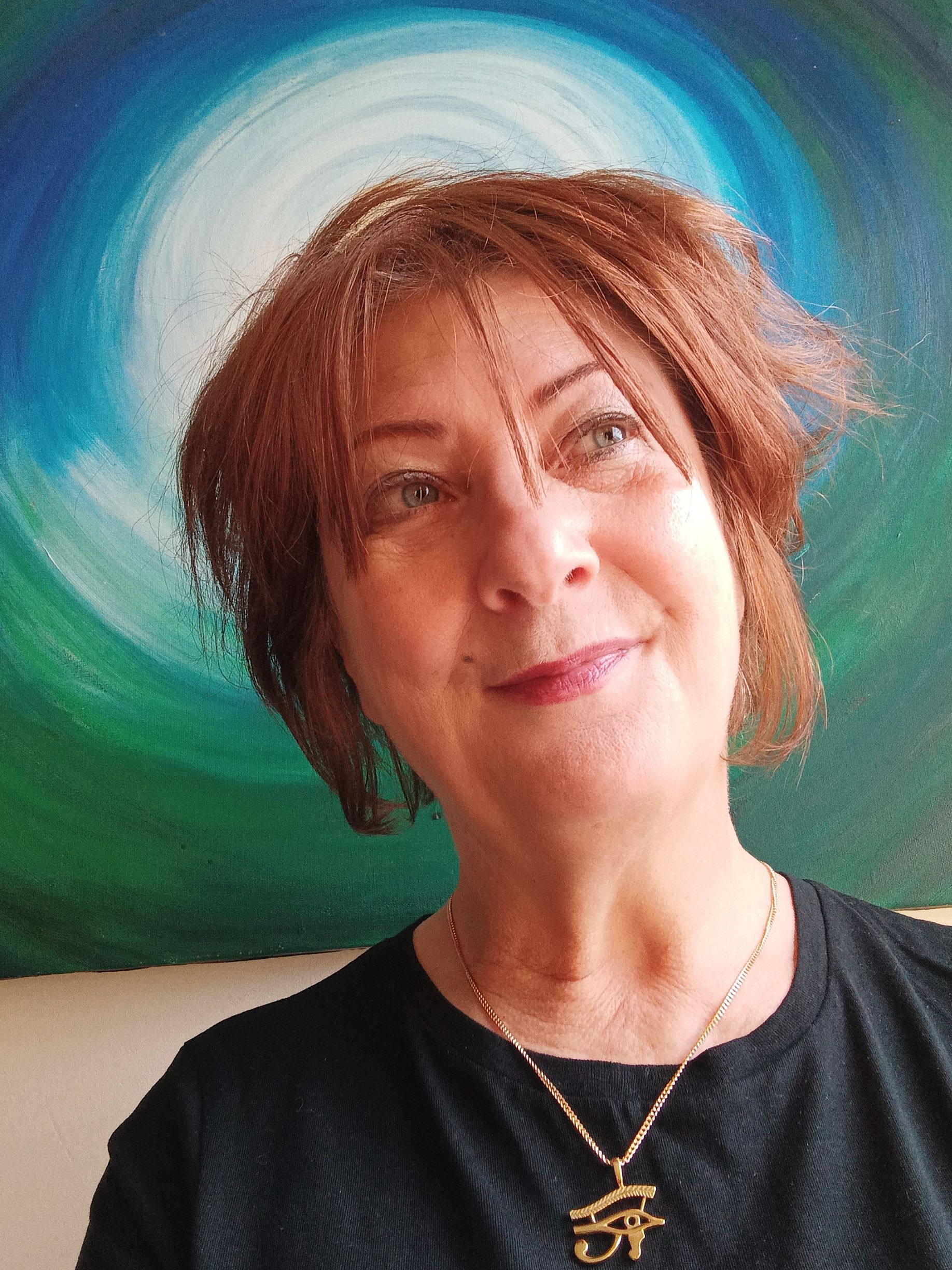 Une femme aux cheveux châtain roux sourit, vêtue d'une chemise noire et d'un collier. Un tableau abstrait bleu et vert orne l'arrière-plan.