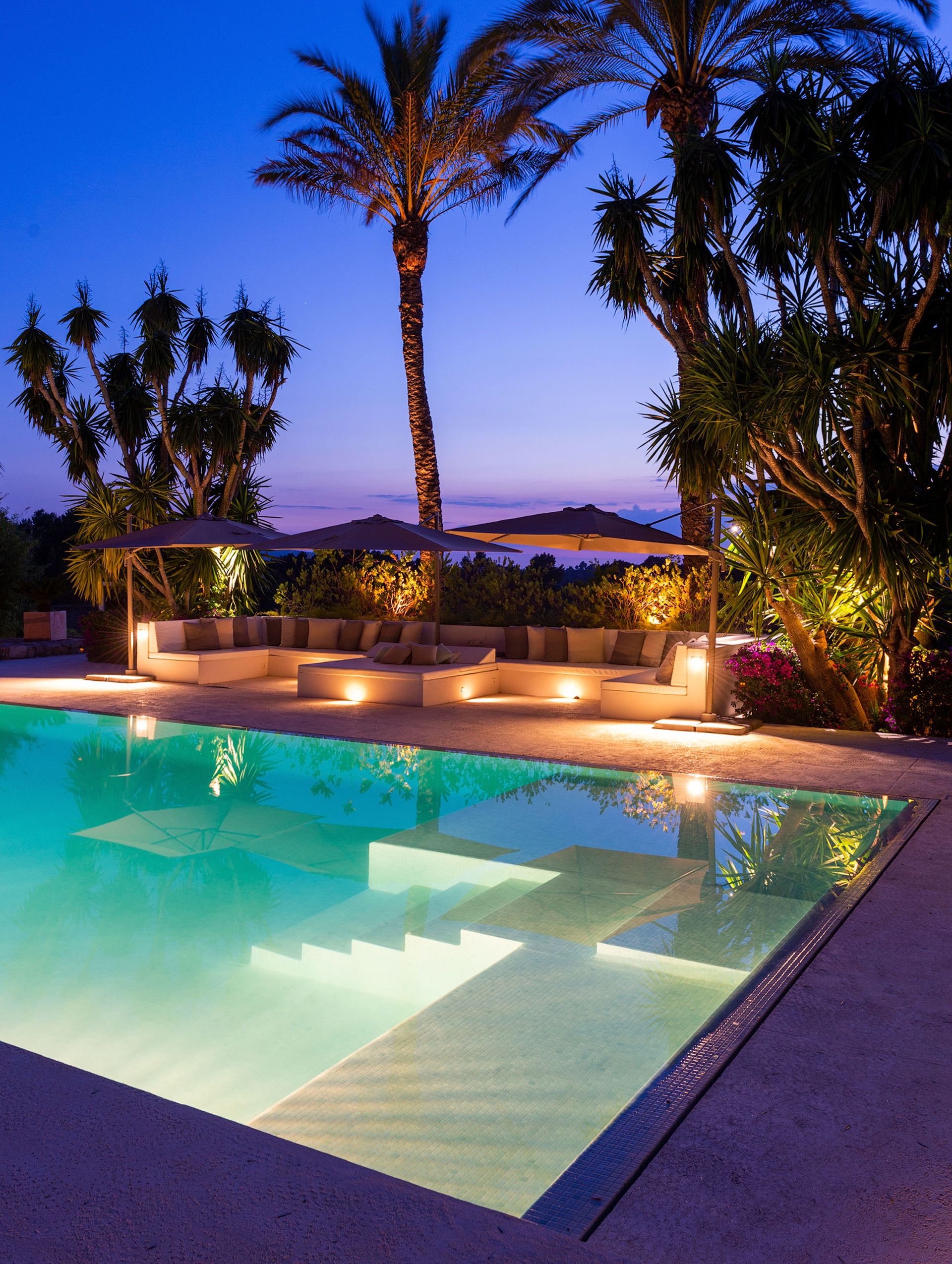 Piscina iluminada al anochecer, con palmeras y tumbonas.