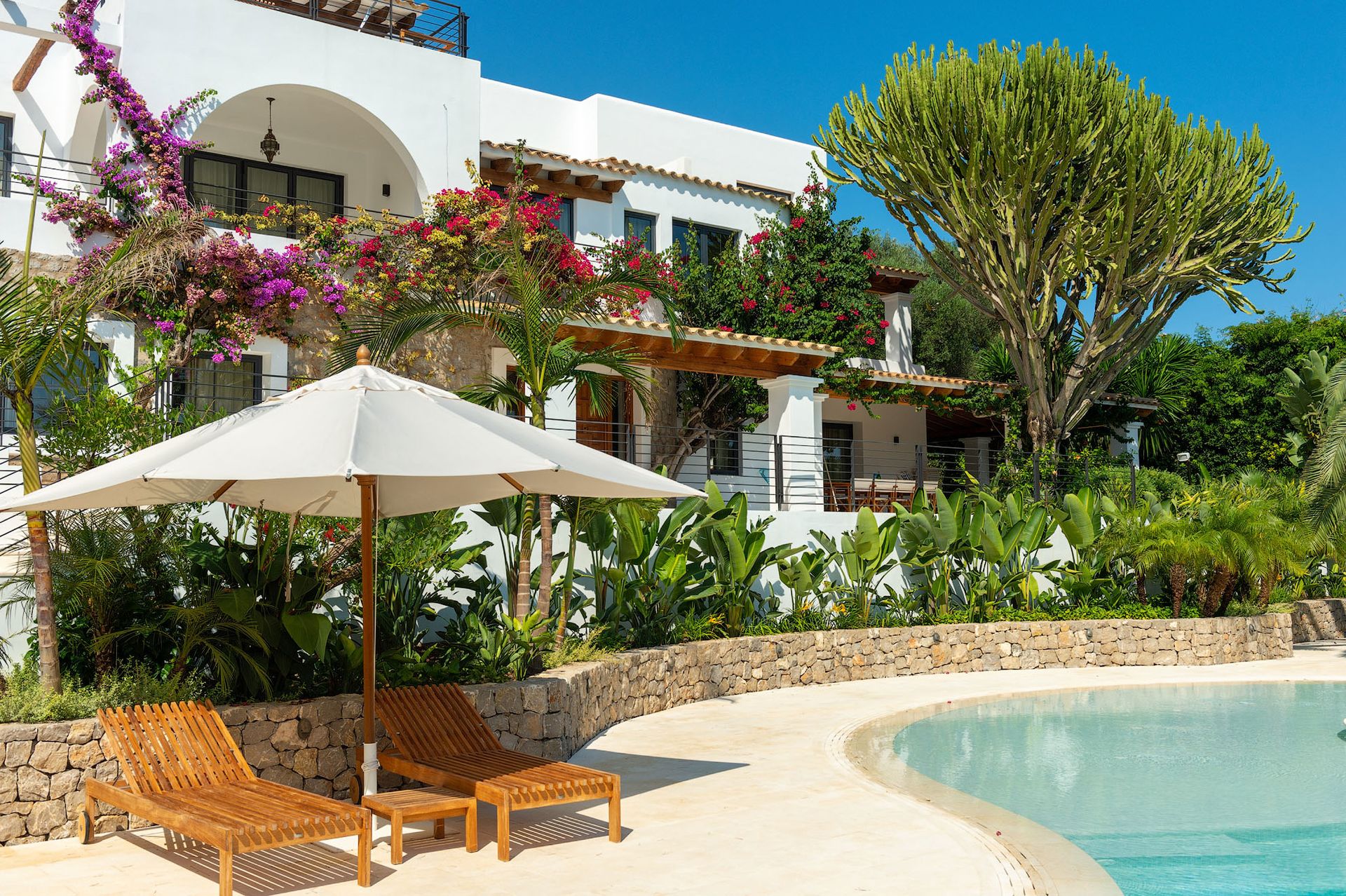Tumbonas bajo una sombrilla blanca junto a una piscina en una villa blanca con exuberante vegetación.