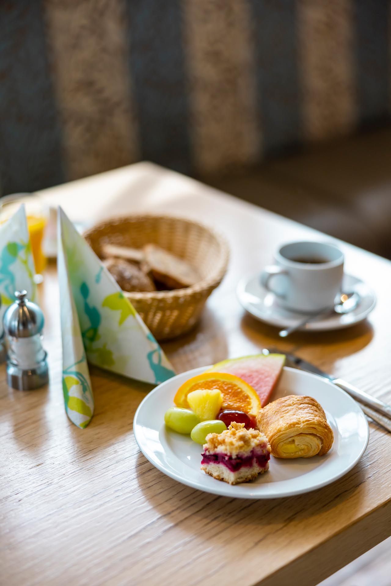 Frühstücksgebäck und Obst auf einem weißen Teller neben einer Tasse Kaffee auf einem Holztisch