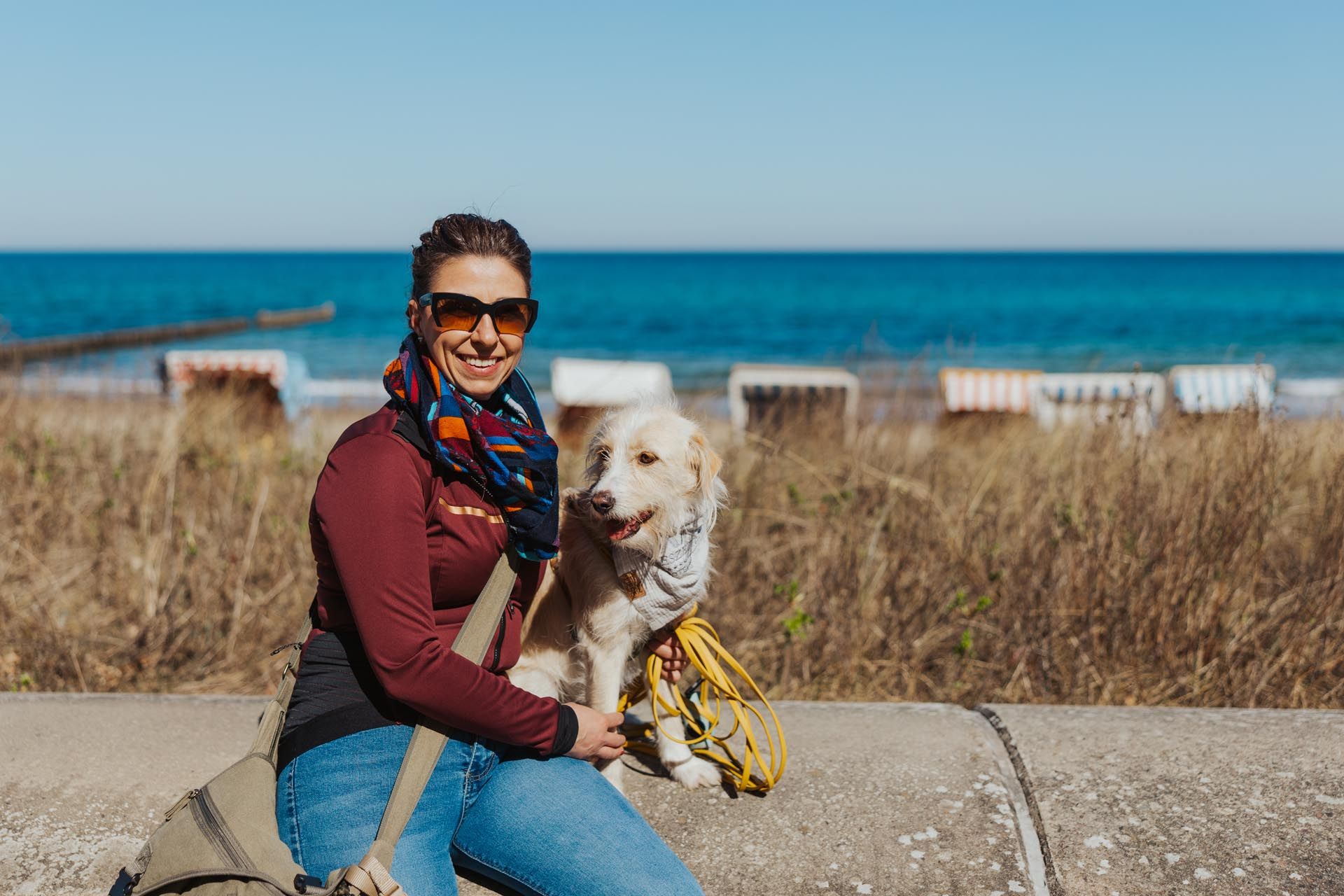 Eine Person kniet mit einem weißen Hund am Strand; im Hintergrund sind das blaue Meer und trockenes Gras zu sehen.