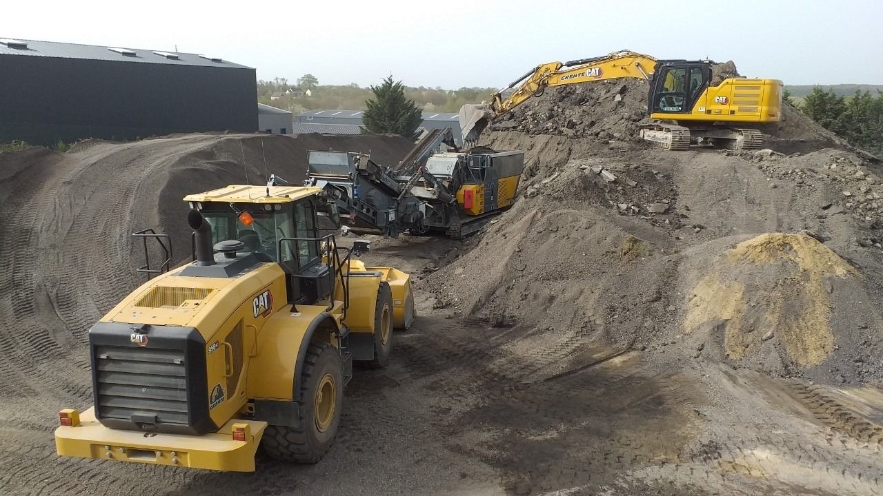 Des engins de chantier jaunes travaillent sur un tas de terre ; un bâtiment et des arbres se trouvent en arrière-plan.