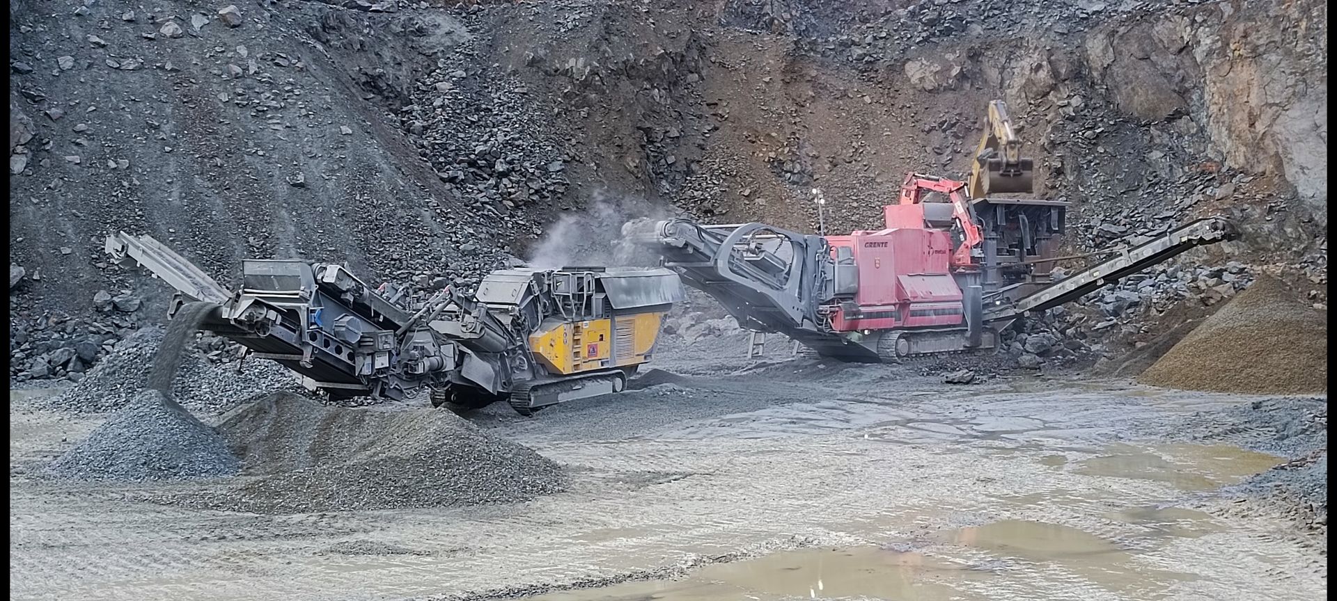 Deux engins de chantier, rouges et jaunes, concassent des roches dans une carrière.