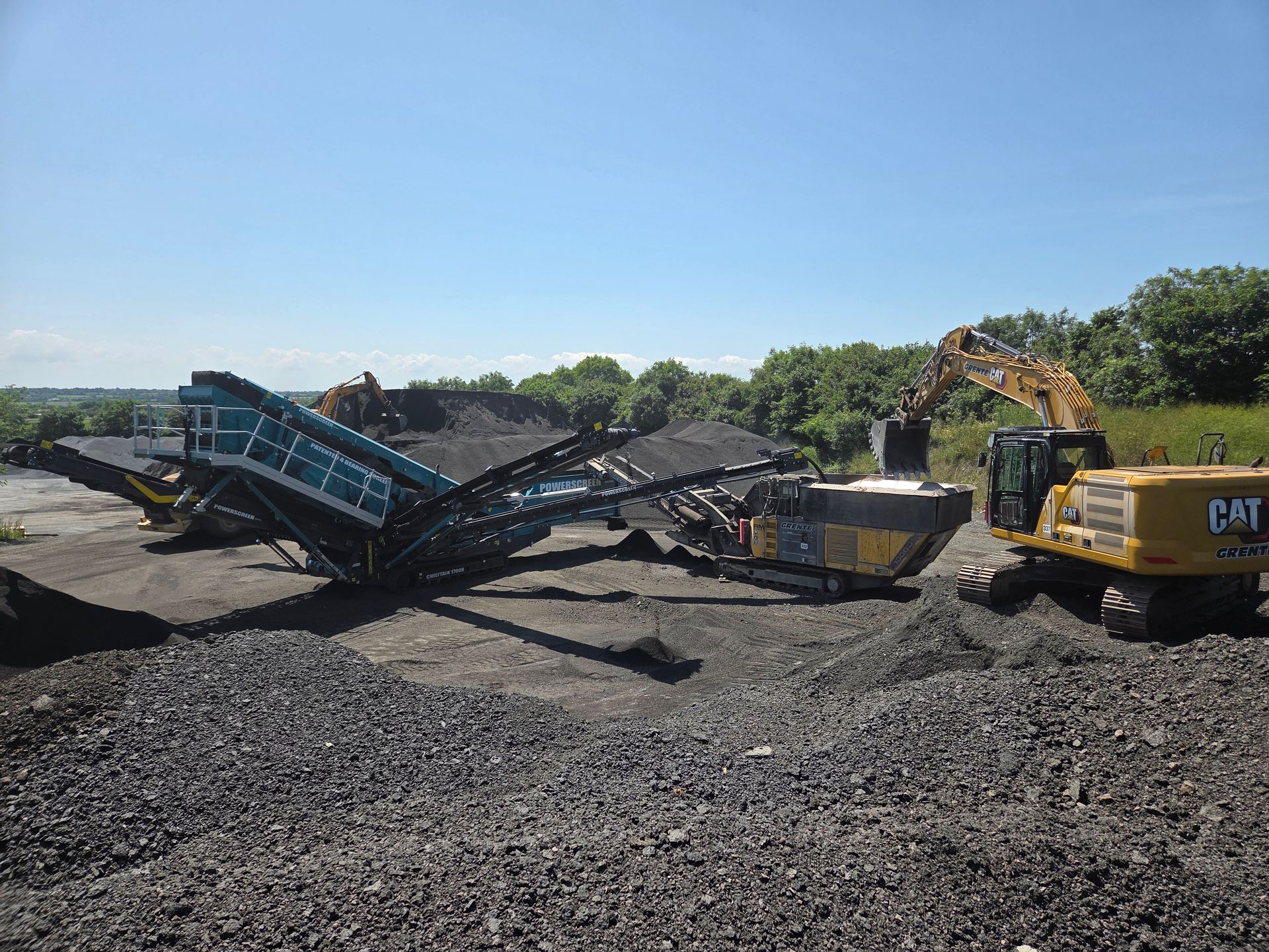 Une excavatrice charge une machine de criblage de roches sur un chantier. Des tas de roches, ciel bleu.