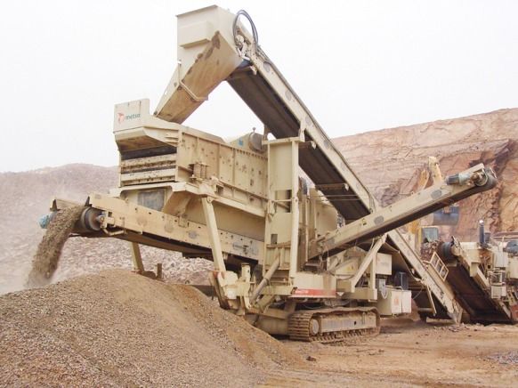 Une imposante machine industrielle de criblage de roches, équipée de convoyeurs à bande, déposant du gravier. Construction métallique beige en extérieur.