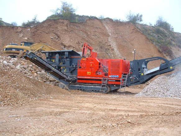 Une machine de concassage de roches rouges dans une carrière, en train de concasser des pierres. Une excavatrice est visible à l'arrière-plan.