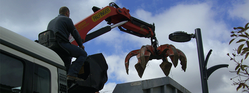 Une grue va prendre des déchets dans une benne près d'un lampadaire.