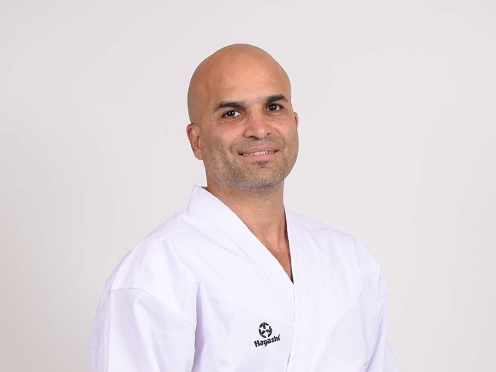 Mann in weißer Karateuniform, lächelnd, vor weißem Hintergrund.