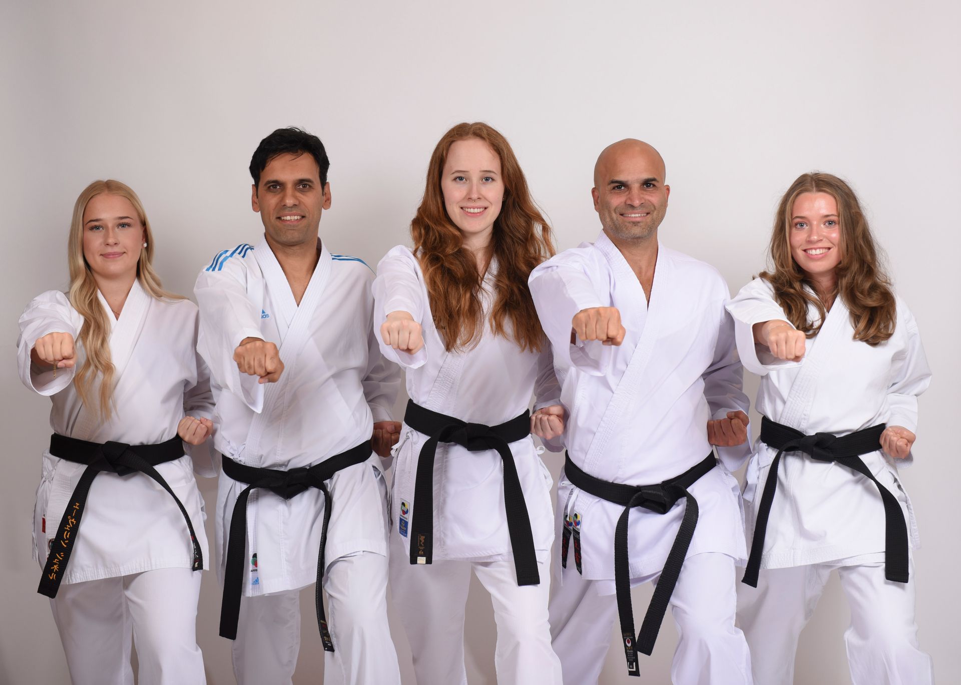 Fünf Personen in weißen Karateuniformen und mit schwarzen Gürteln stoßen vor einem weißen Hintergrund ihre Fäuste nach vorne.