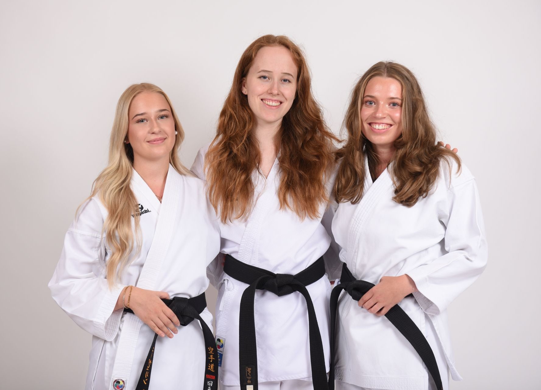 Drei Frauen in Karateuniformen, schwarze Gürtel, lächelnd vor einem weißen Hintergrund.