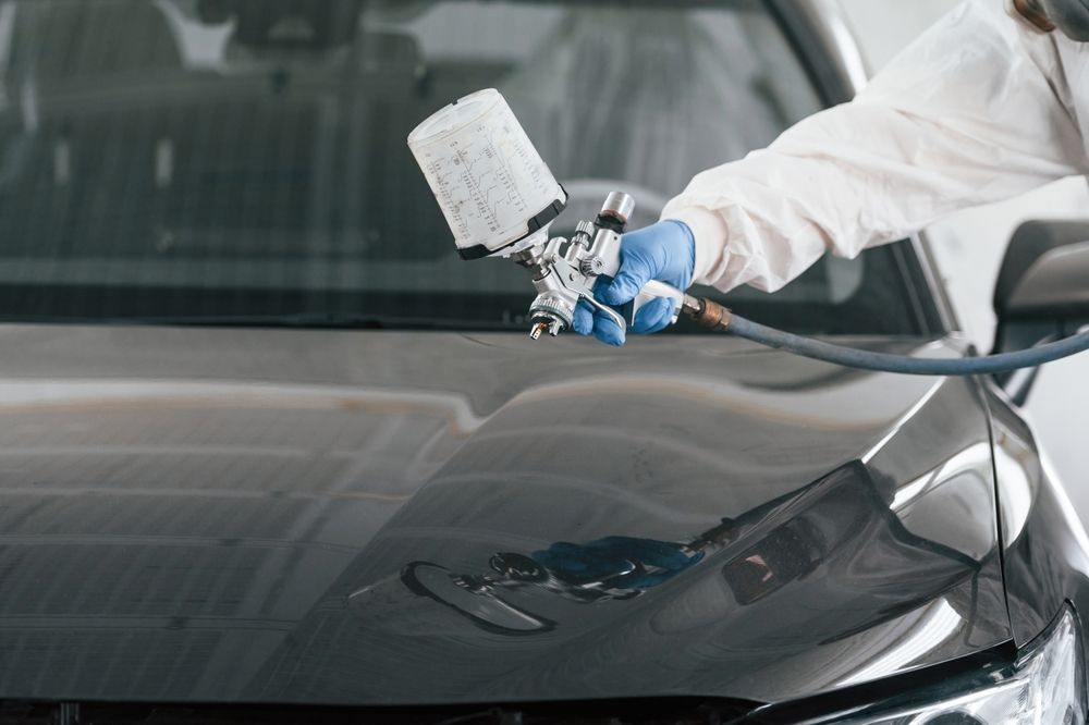 Una persona con equipo de protección utiliza una pistola de pintura en aerosol para aplicar un acabado brillante al capó de un coche negro.