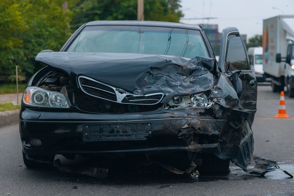 Un sedán negro gravemente dañado permanece en medio de la carretera tras una colisión, con un cono de tráfico cerca.