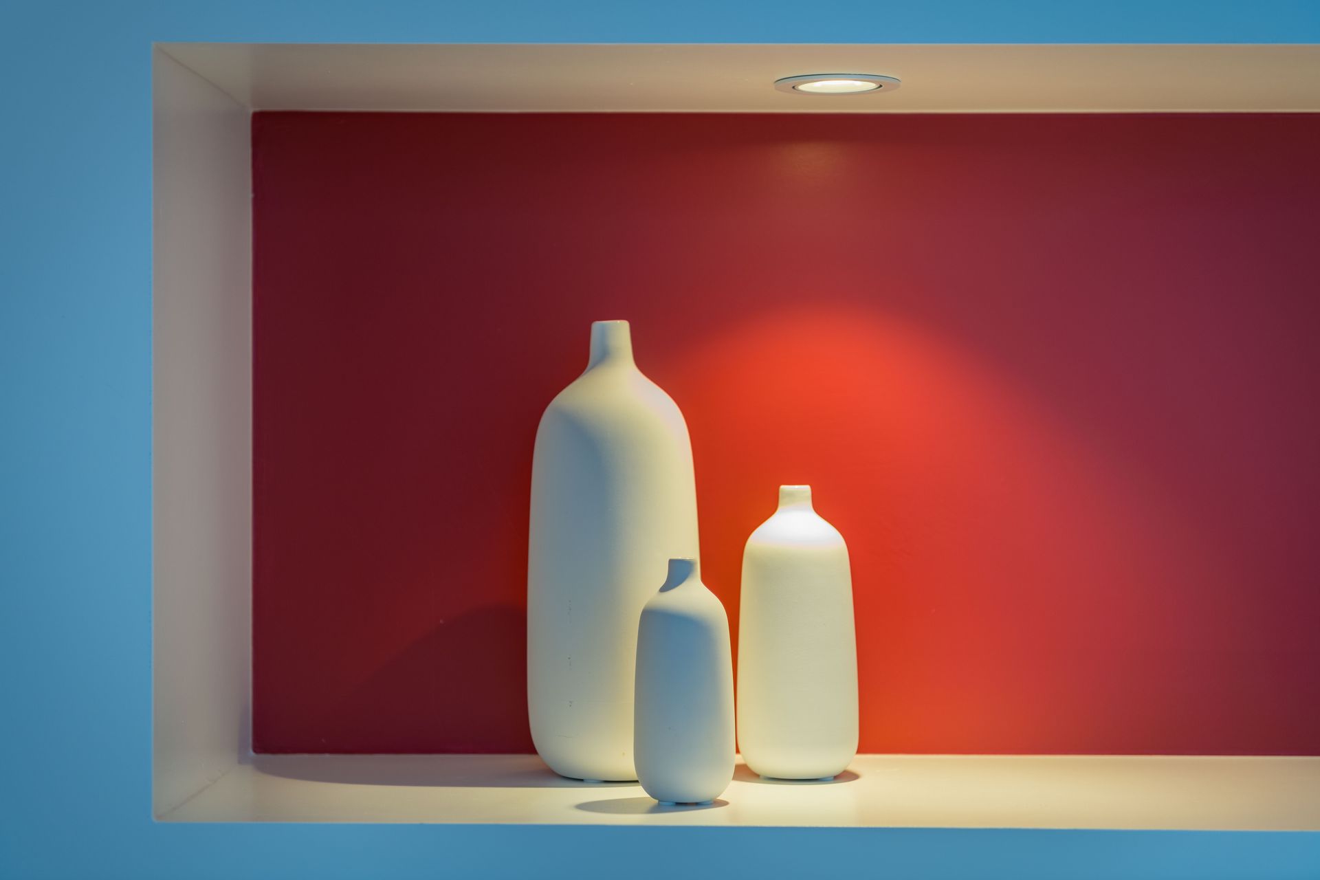 Three white vases are on a shelf in front of a red wall