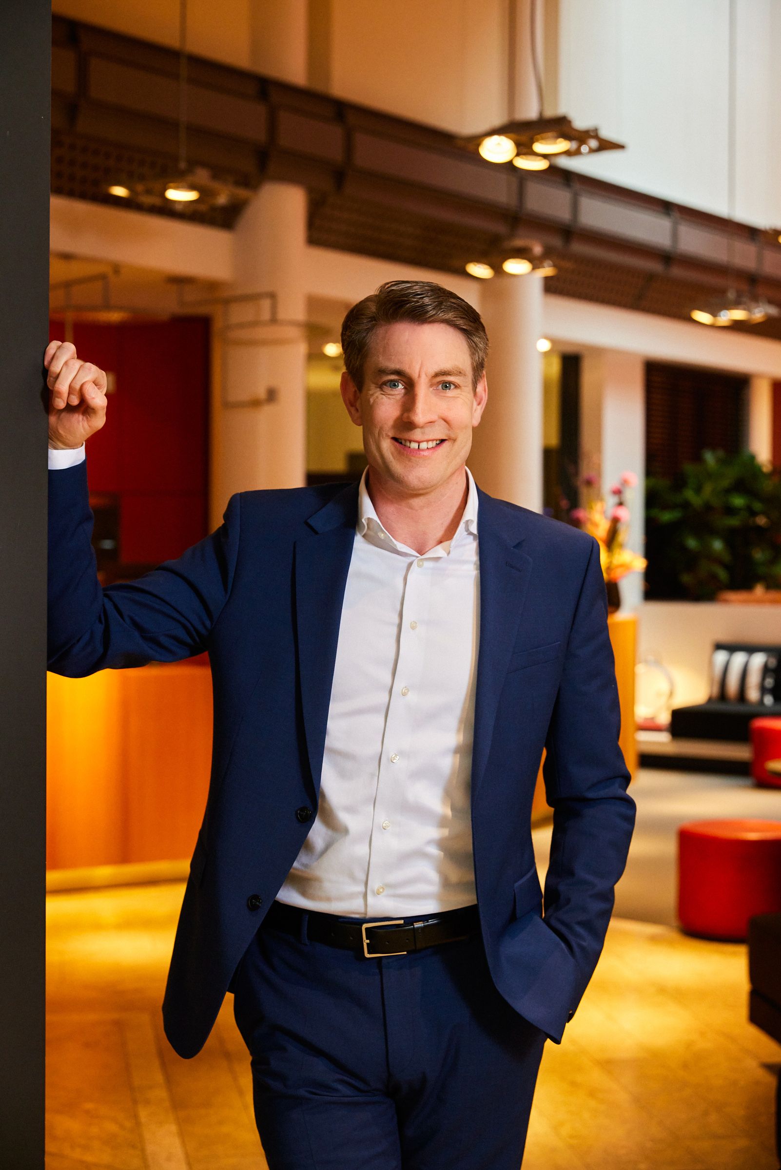 A man in a suit and white shirt is standing in a lobby with his hands in his pockets.