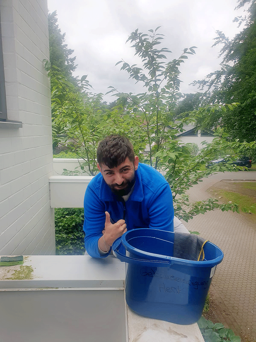 Ein Mann in einer blauen Jacke sitzt auf einem Balkon neben einem blauen Eimer und lächelt.