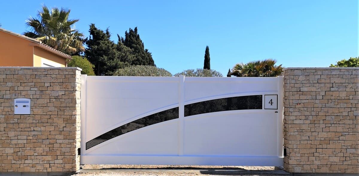 Portail blanc orné de motifs noirs entre des murs de briques, ciel bleu en arrière-plan.