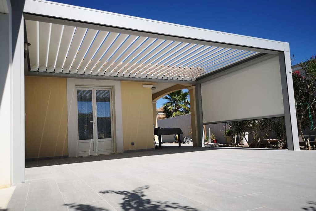 Terrasse extérieure moderne avec toit blanc rétractable, bâtiment beige et sol gris clair, sur fond de ciel bleu.
