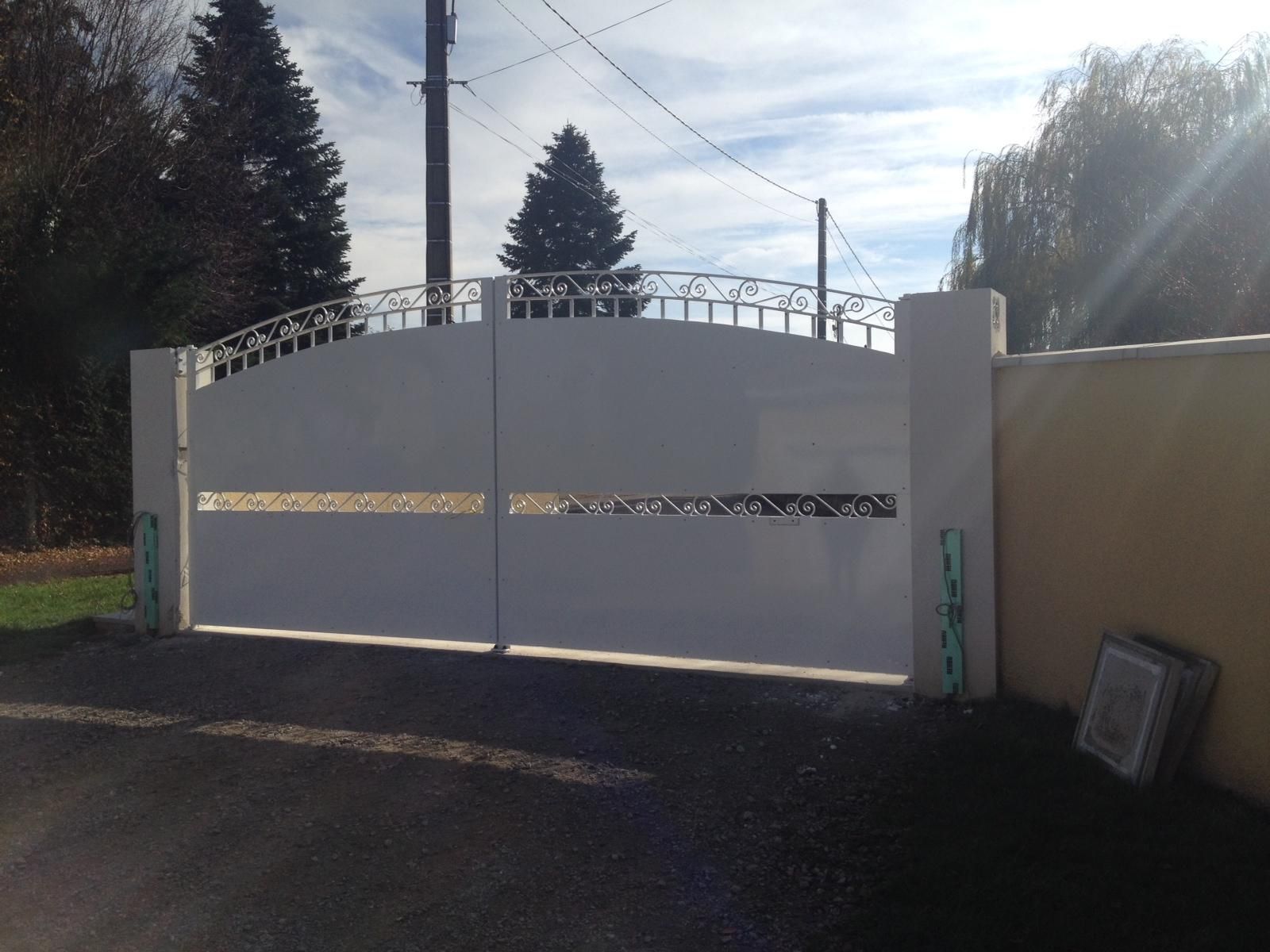 Portail en métal blanc orné d'un élément décoratif sur une allée pavée baignée de soleil, avec des arbres et des poteaux électriques en arrière-plan.