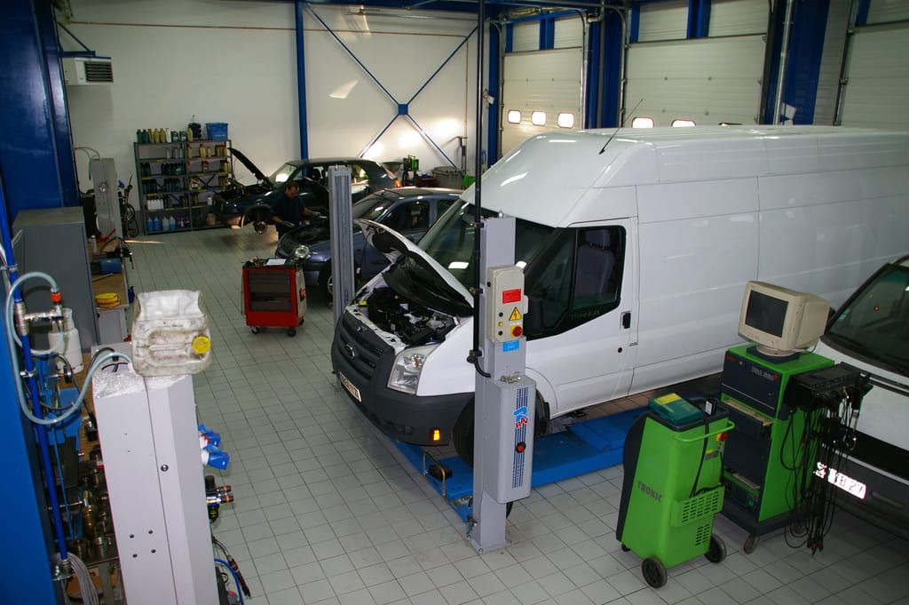 Fourgonnette blanche dans un garage de réparation automobile, capot ouvert et entourée d'outils et d'équipements.