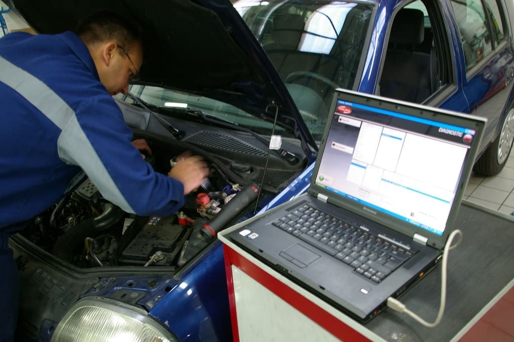 Un mécanicien travaille sous le capot d'une voiture bleue, à côté d'un ordinateur portable affichant un logiciel de diagnostic.