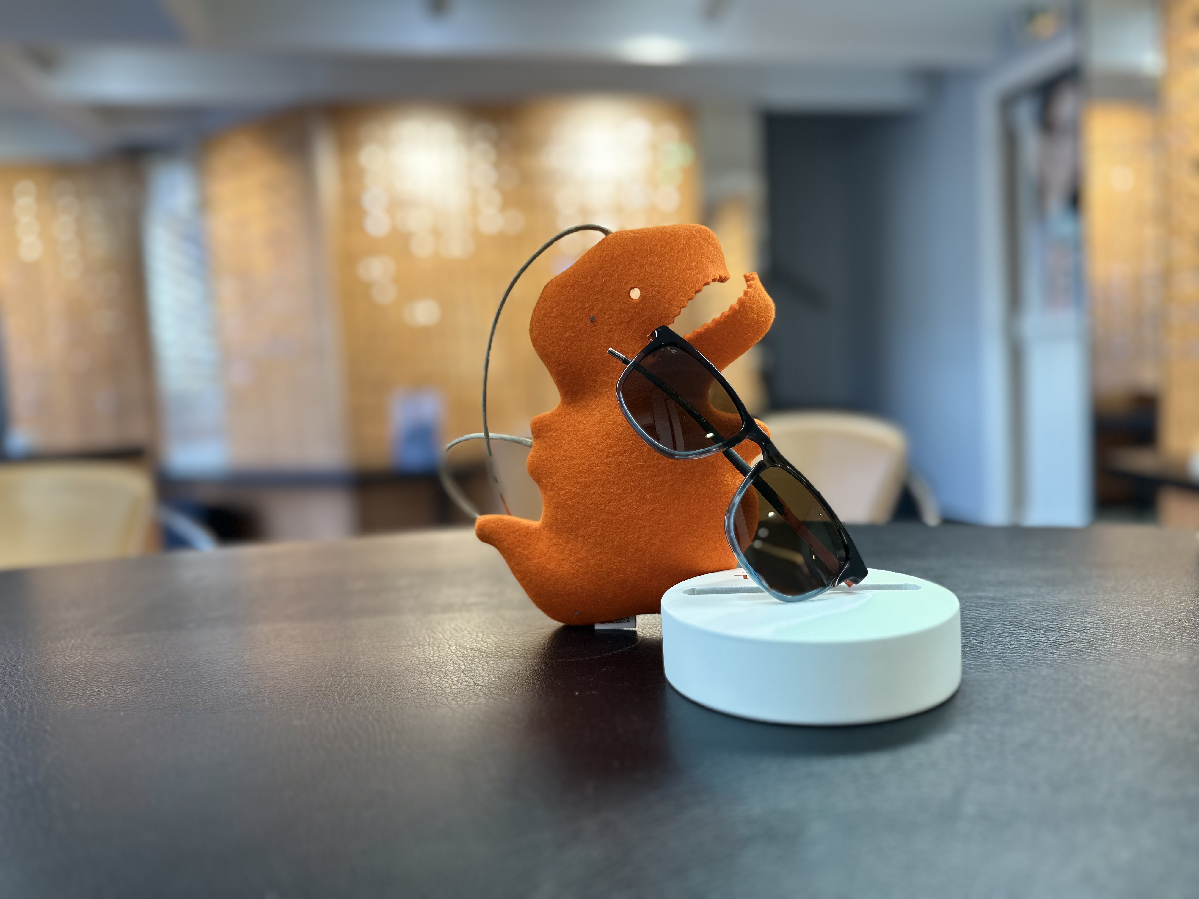 Jouet dinosaure vert tenant des lunettes de soleil orange sur un socle blanc, table noire.