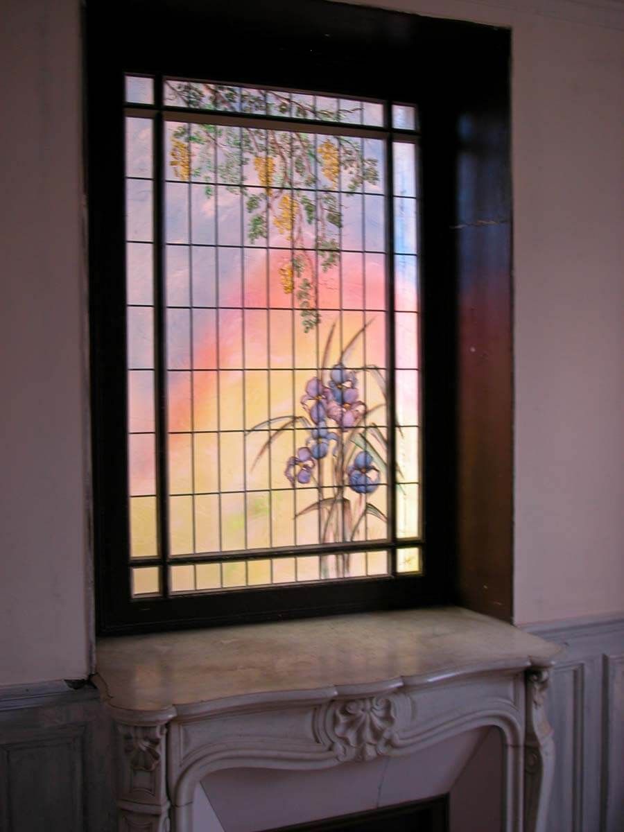 Vitrail à motifs floraux, encadré d'un cadre sombre au-dessus d'une cheminée blanche. Fond couleur ciel.