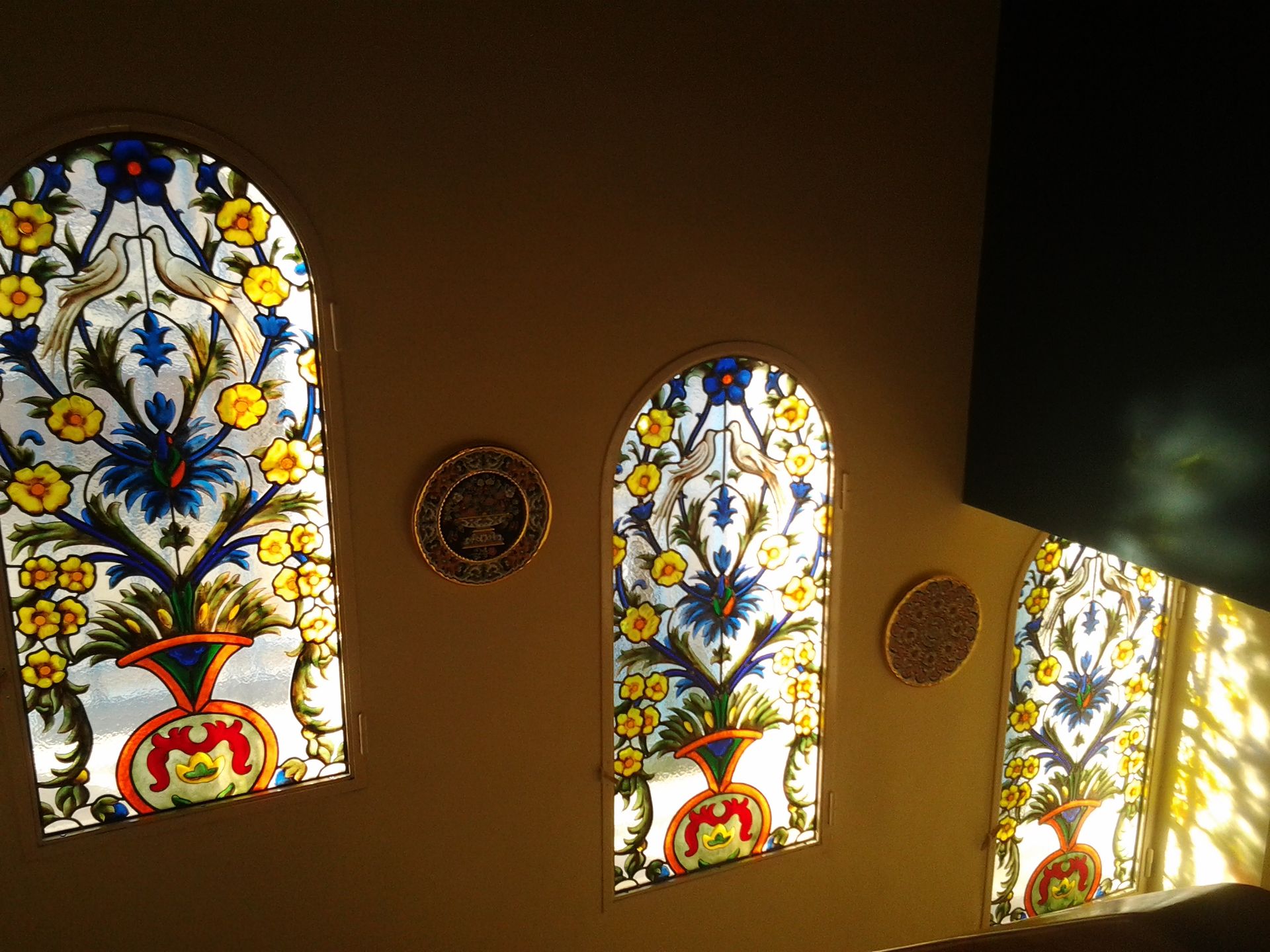 Trois vitraux cintrées à motifs floraux, jaunes et bleues, encastrées dans un mur brun clair, baignées de soleil.