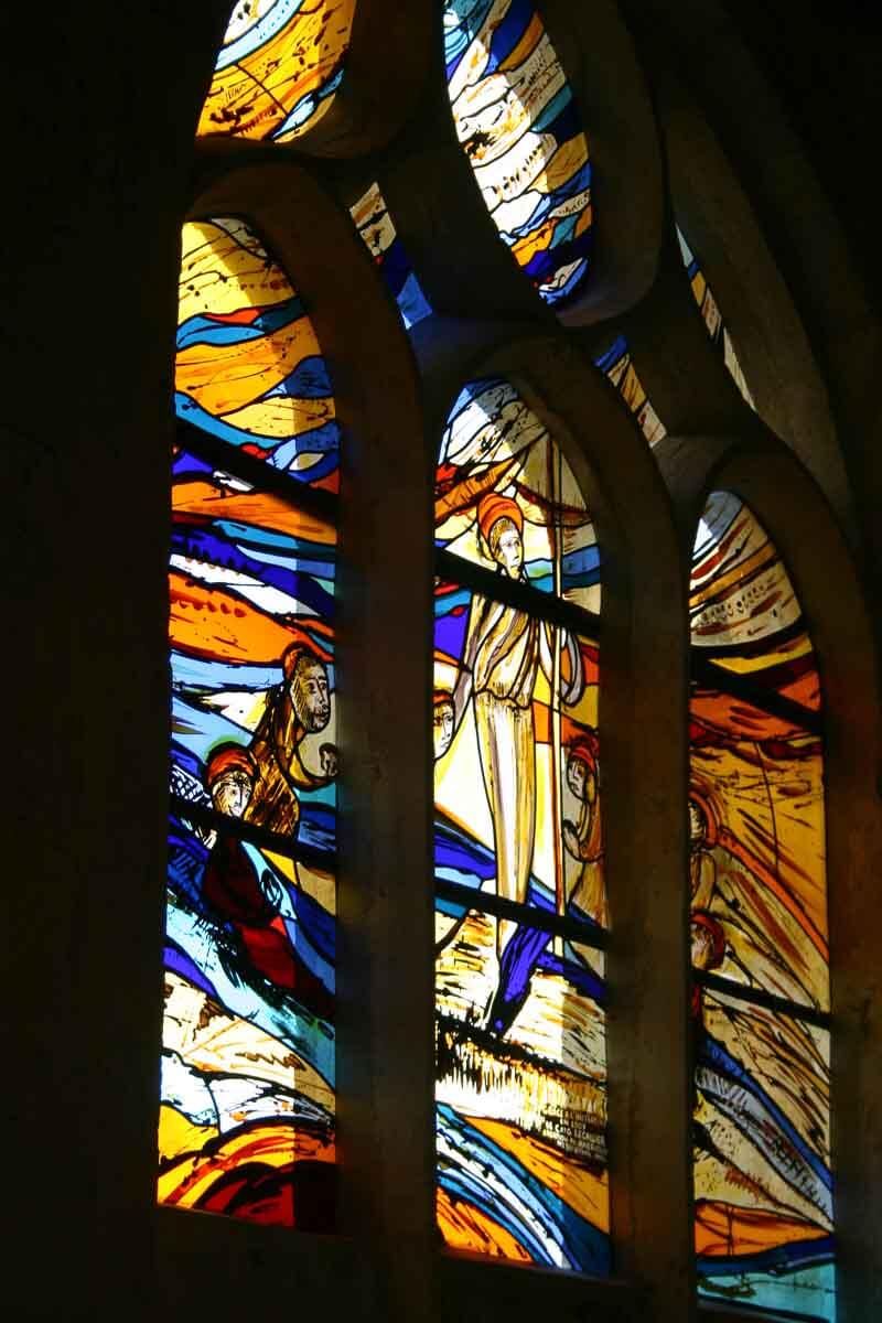 Vitrail orné de formes abstraites bleues, oranges et jaunes, et de silhouettes suggérées dans un cadre arqué.