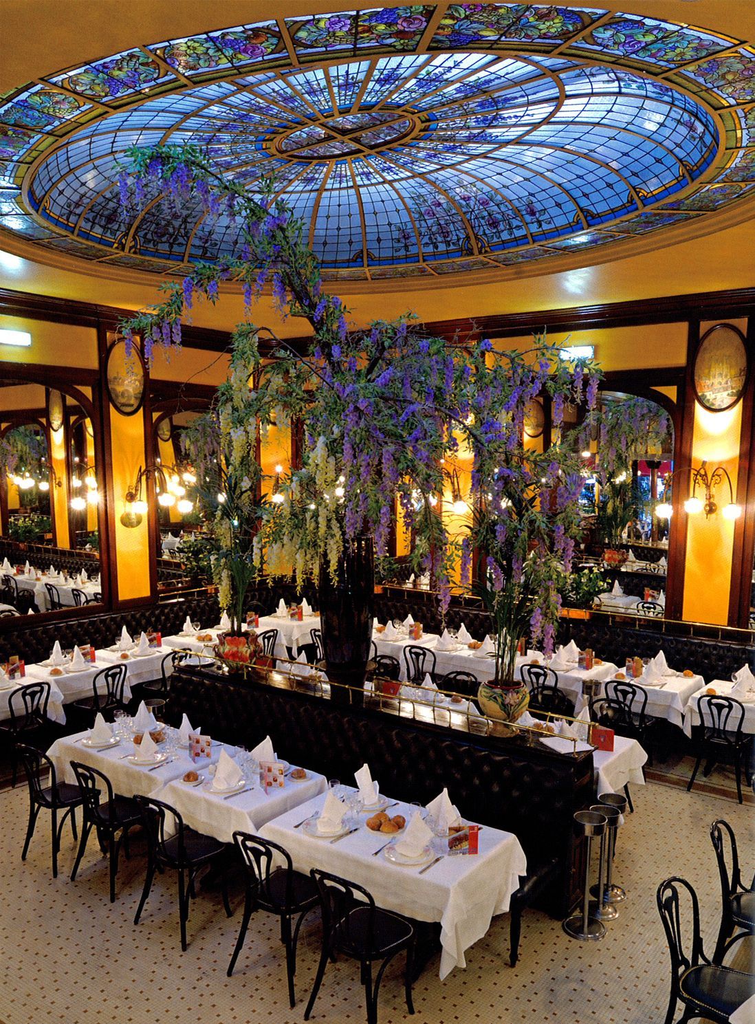 Intérieur élégant d'un restaurant parisien, avec un plafond en vitrail et des tables dressées pour le dîner.