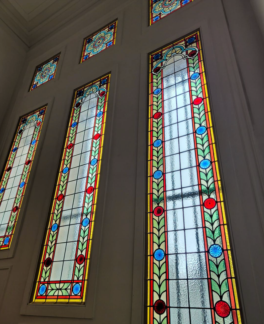 De longues fenêtres en vitrail richement décorées, à bordure colorée, se détachent sur un mur gris clair.