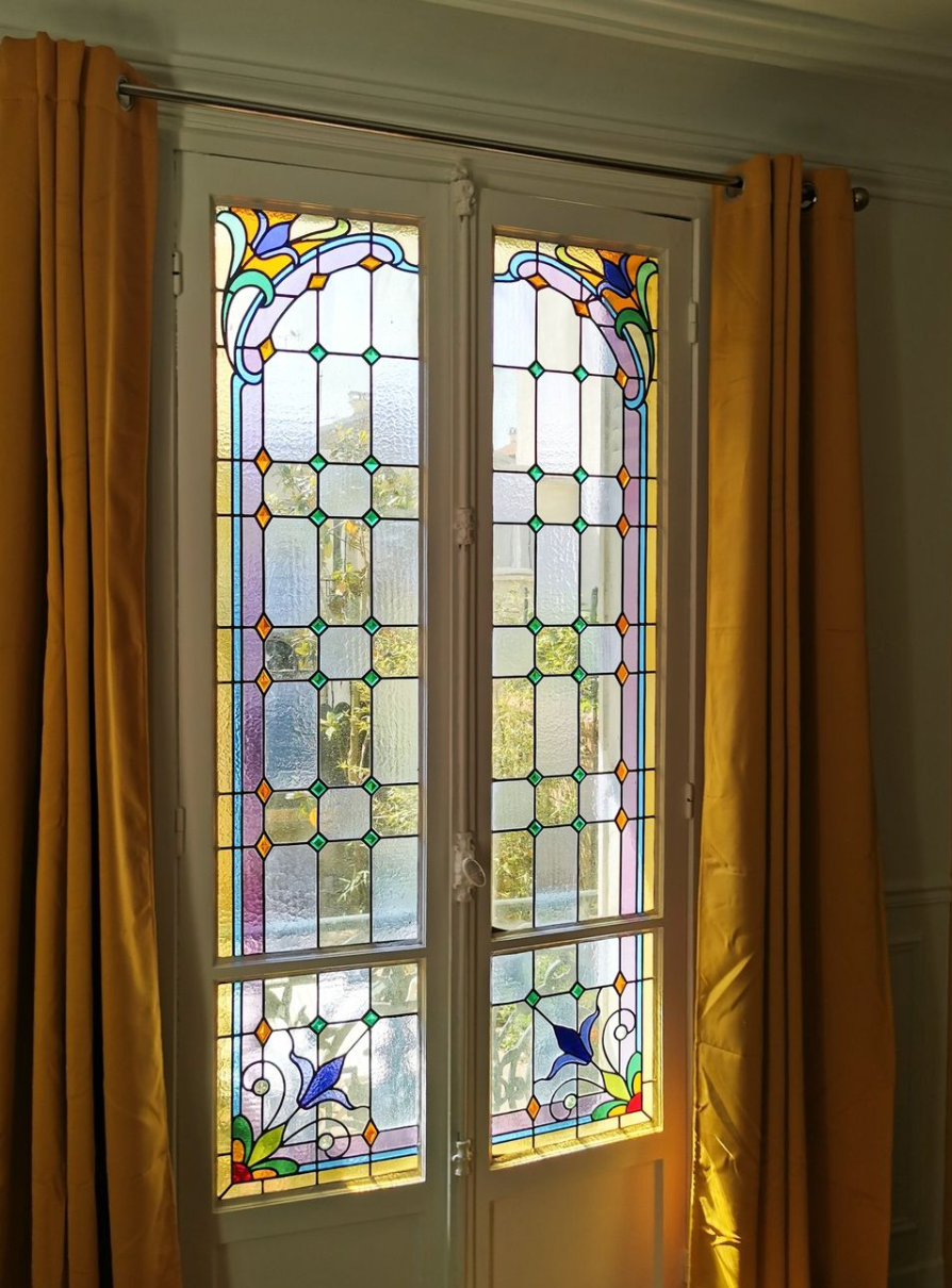 Vitrail aux motifs complexes, encadré de moulures blanches et flanqué de rideaux jaunes.