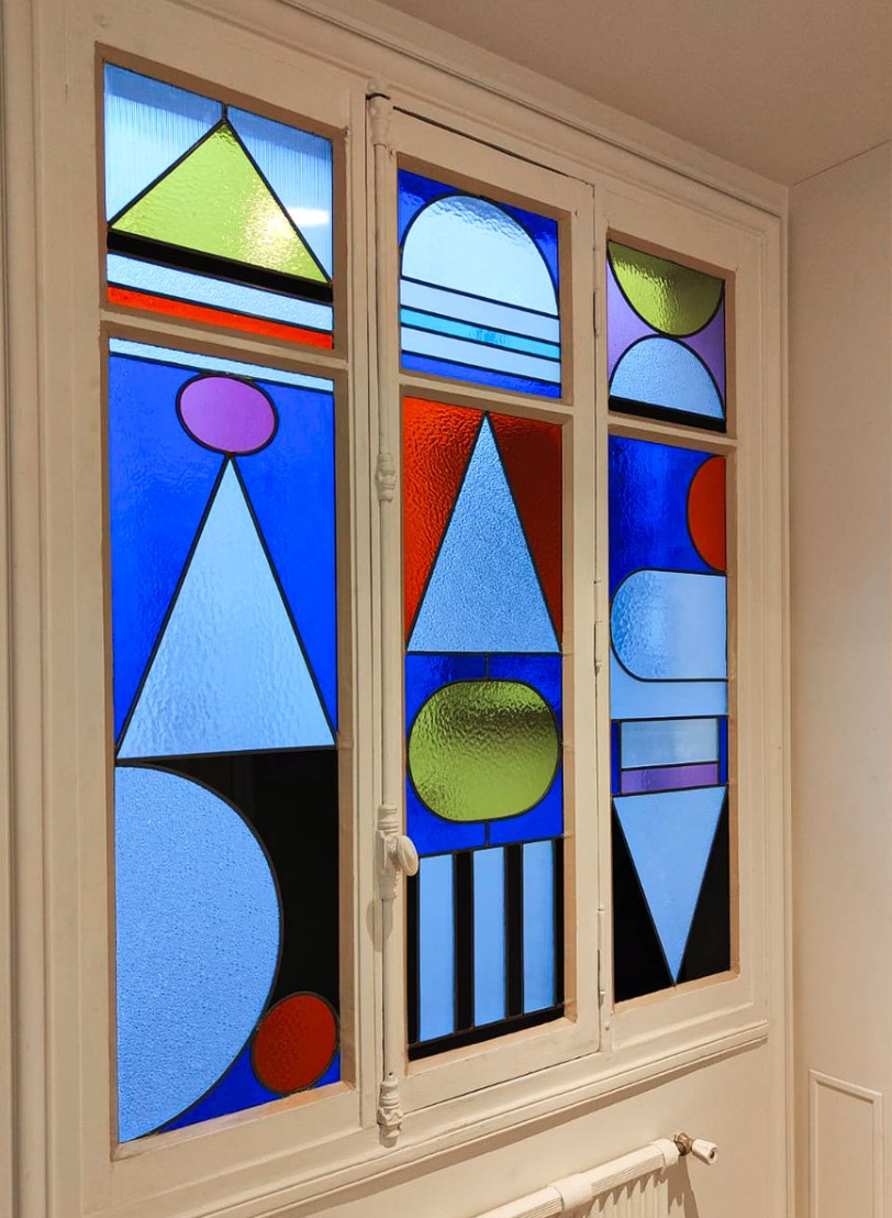 Vitrail géométrique aux formes bleues, rouges, vertes et violettes. Cadre blanc.