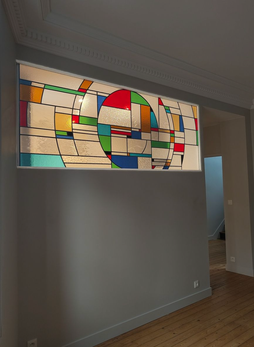 Vitrail coloré dans un cadre blanc au-dessus d'un mur gris. Intérieur avec parquet et porte d'entrée.