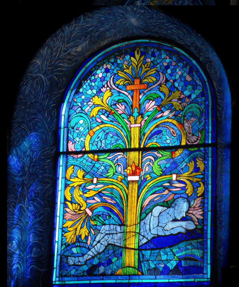 Vitrail représentant un arbre avec une croix et une figure agenouillée dans des tons bleus, jaunes et orange.