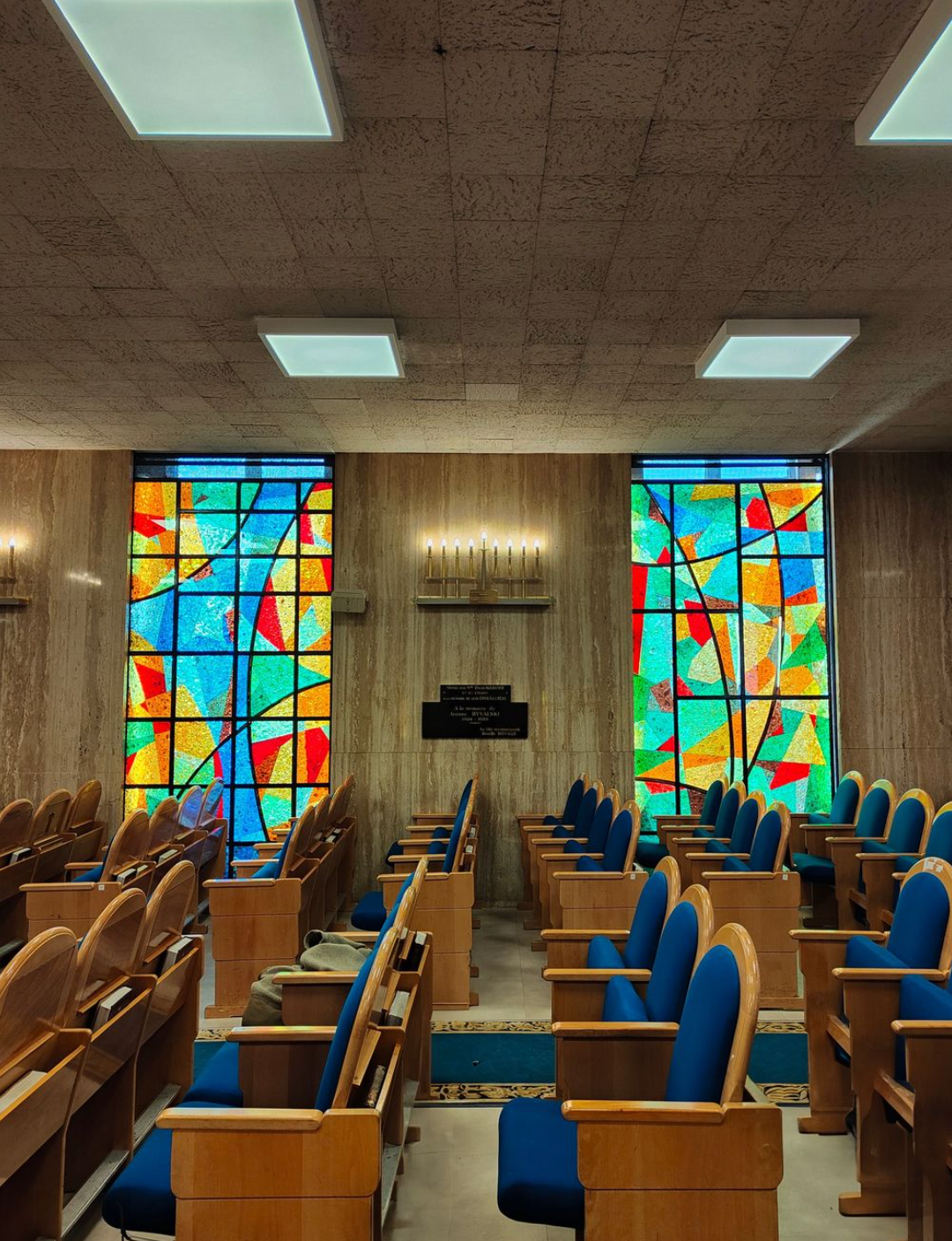 Intérieur d'une synagogue avec bancs en bois, chaises bleues et vitraux.