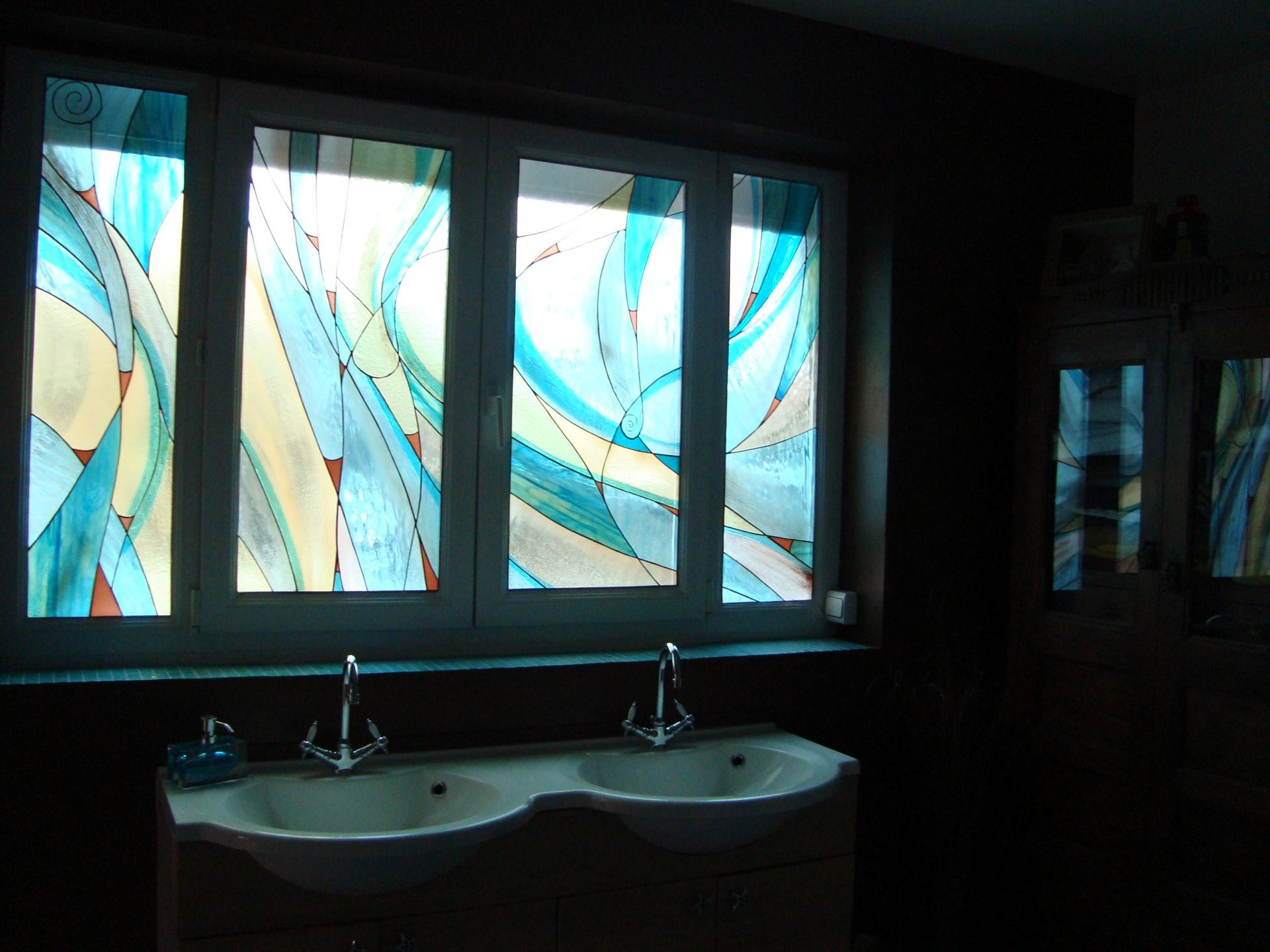 Salle de bains avec un double lavabo sous une fenêtre ornée de vitraux abstraits dans les tons bleus, jaunes et blancs.