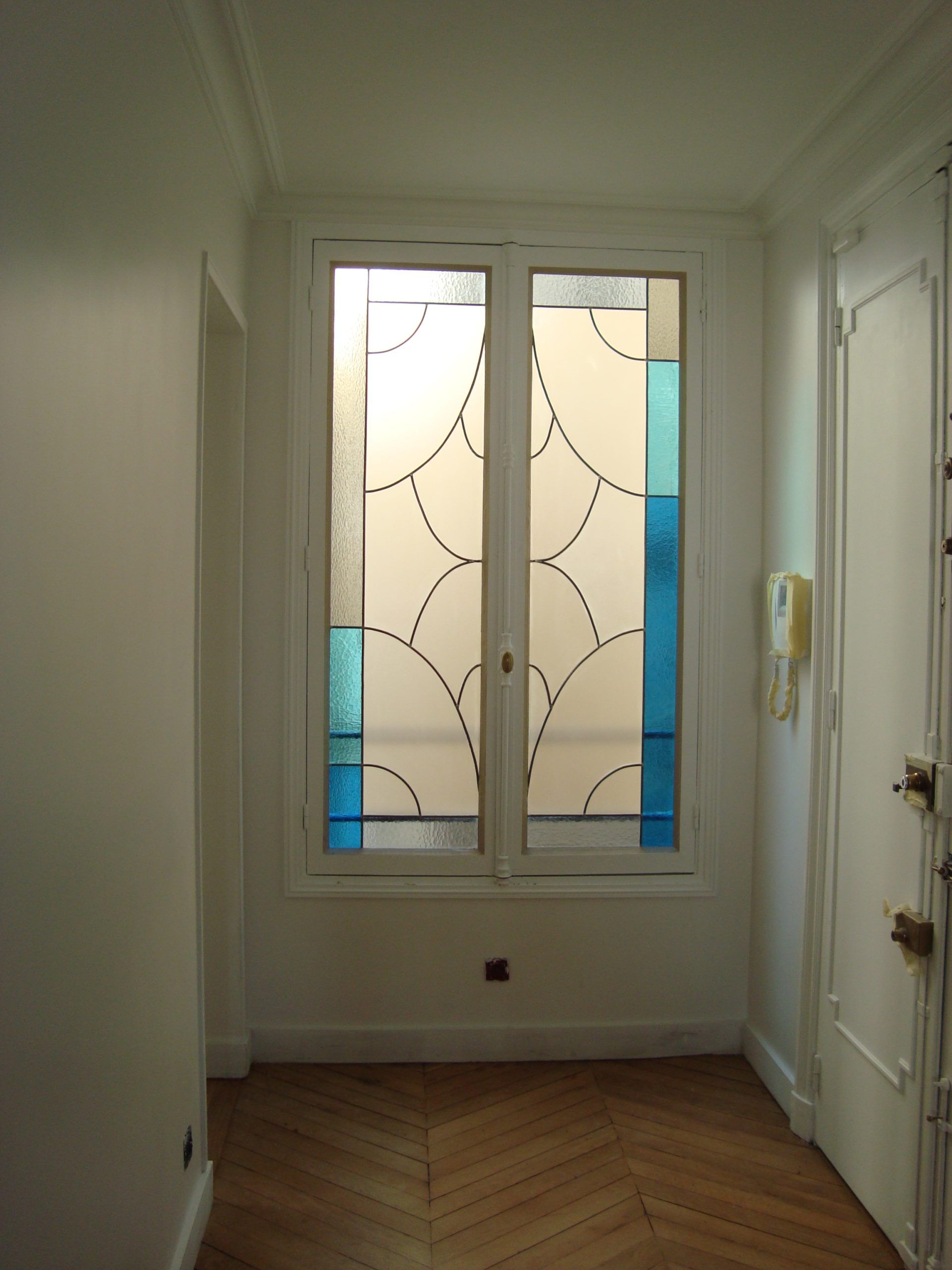 Couloir aux murs blancs, avec un vitrail aux accents bleus et un parquet.