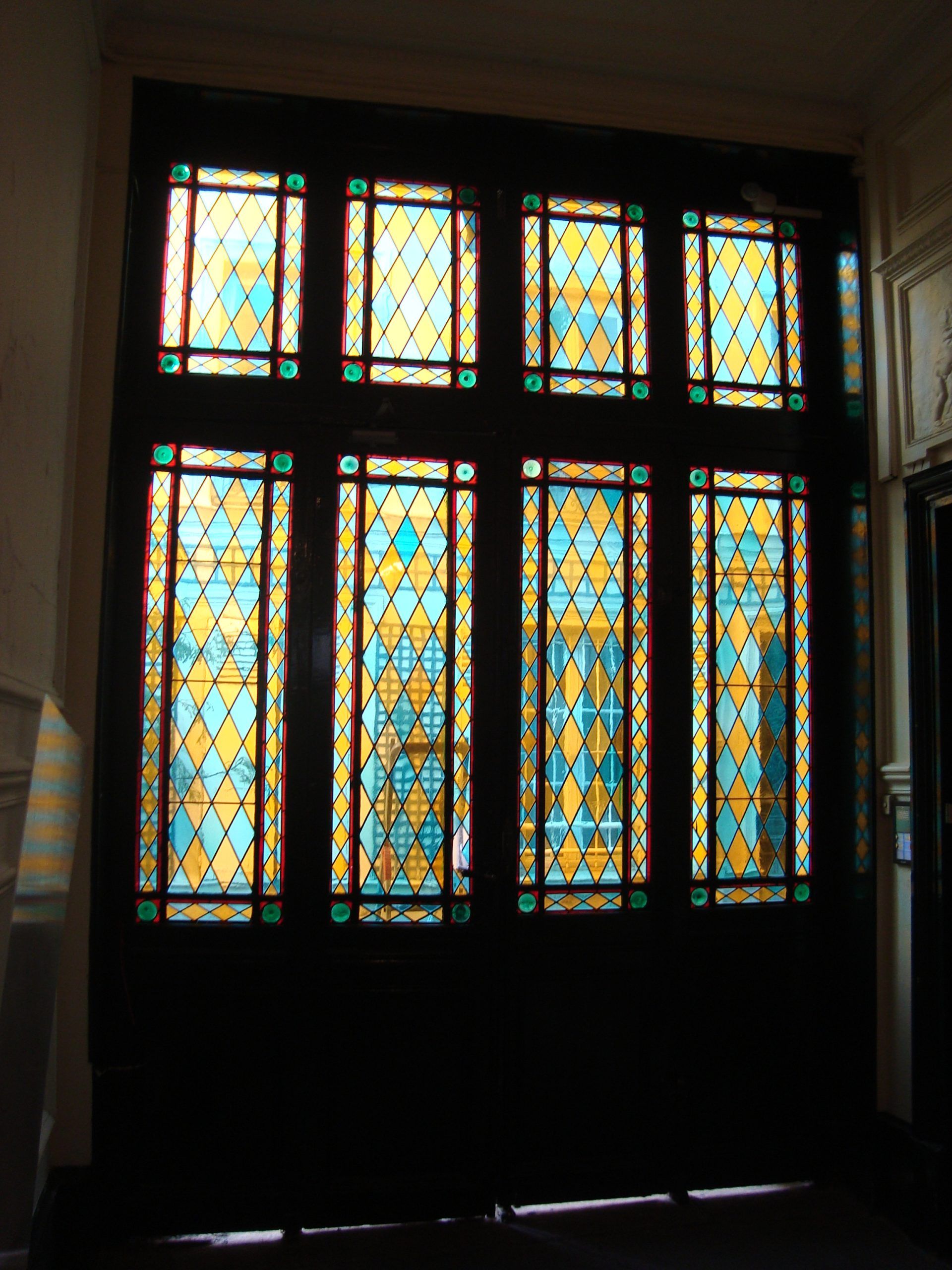 Vitrail orné de losanges turquoise, or et verts, encadré de bois sombre. La lumière du soleil le traverse.