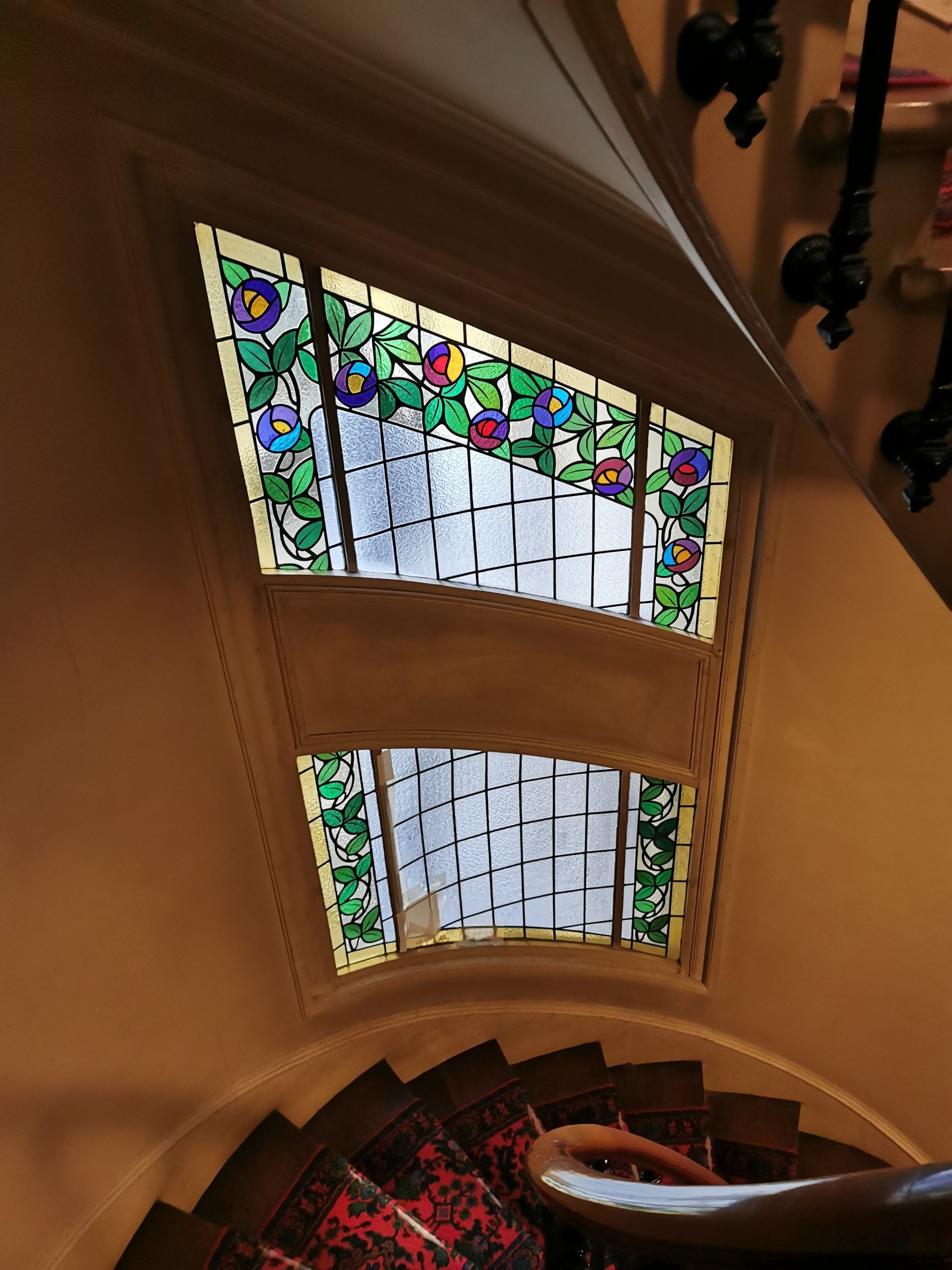 Vitrail à motifs floraux, au-dessus d'un escalier courbe recouvert d'un tapis rouge à motifs.