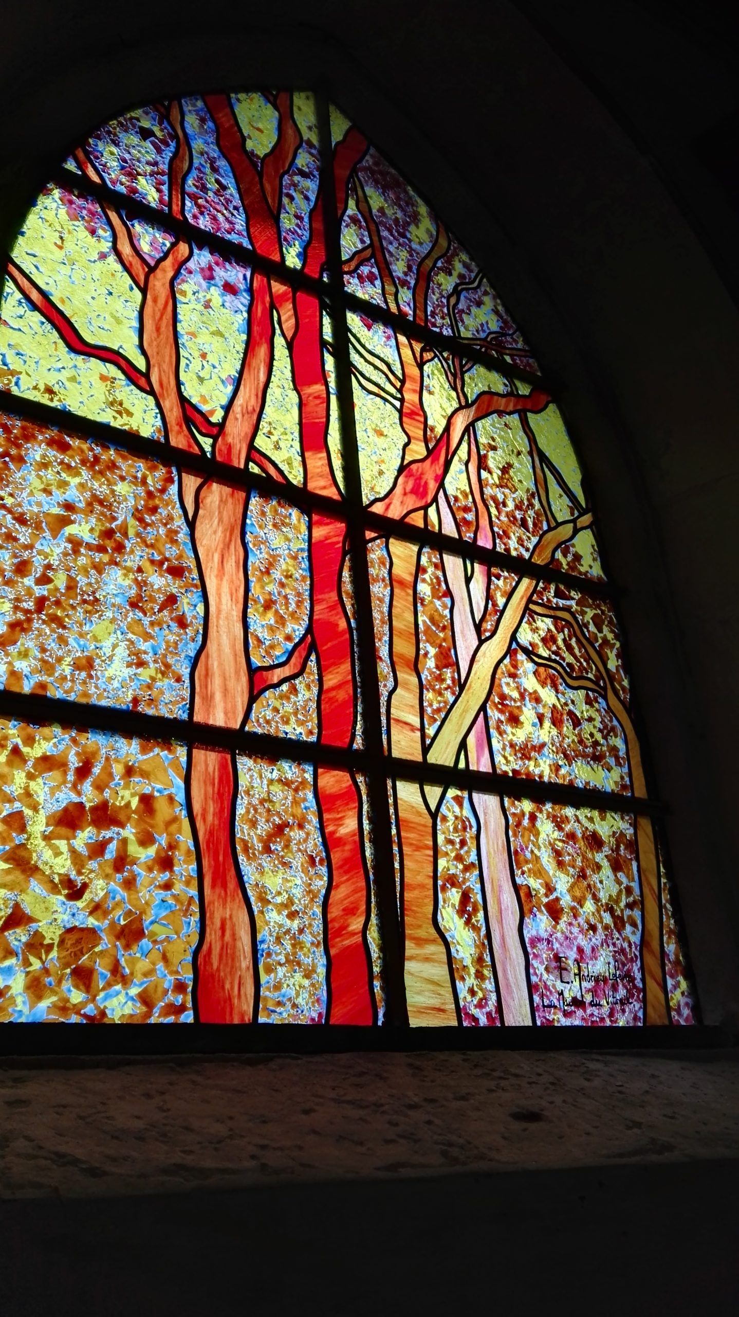 Vitrail représentant des arbres aux troncs rouges et orange, sur un fond de mosaïque aux couleurs d'automne.