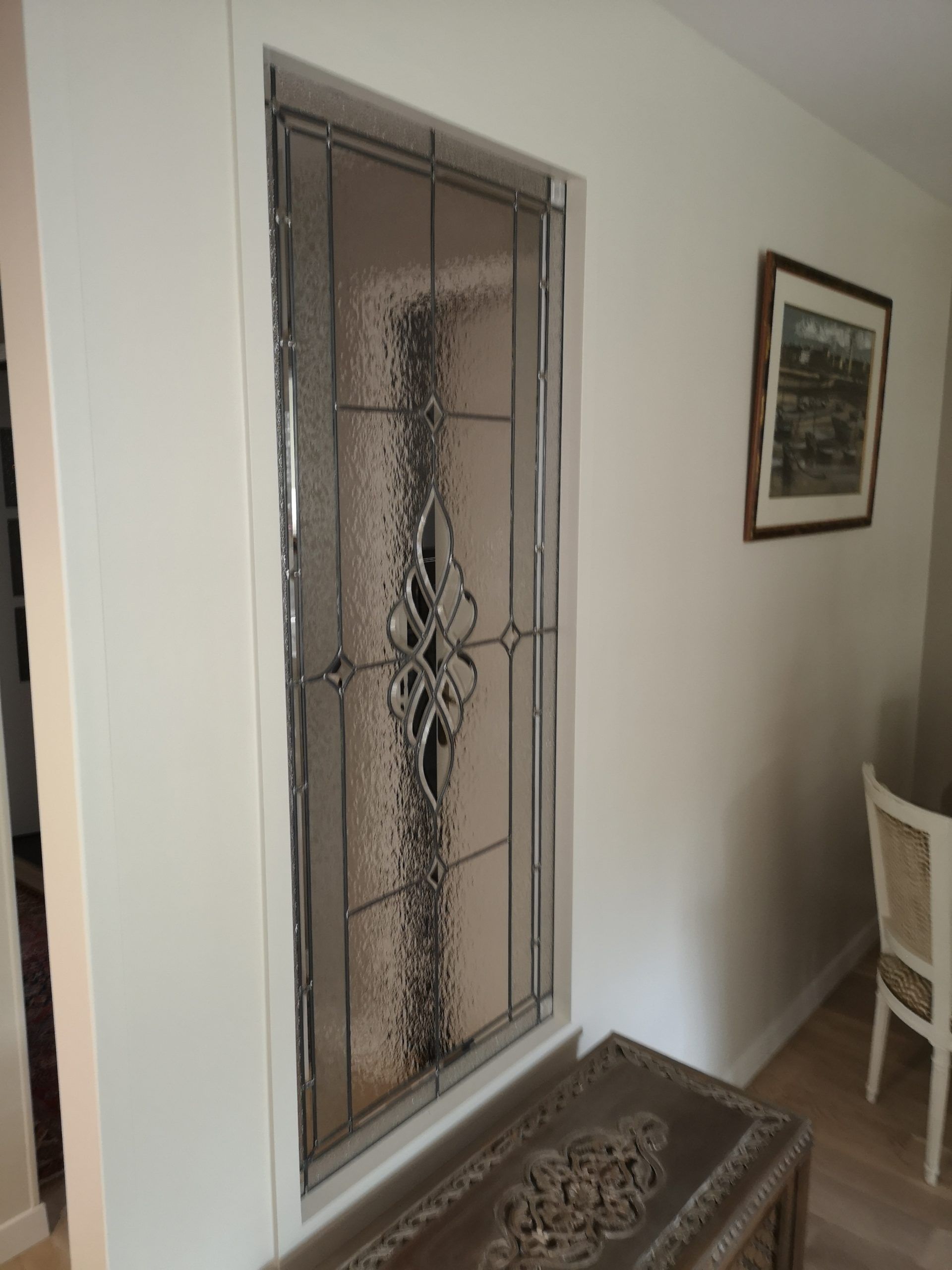 Vitrail encadré dans un mur blanc, orné de motifs décoratifs. Un tableau encadré est accroché à droite.