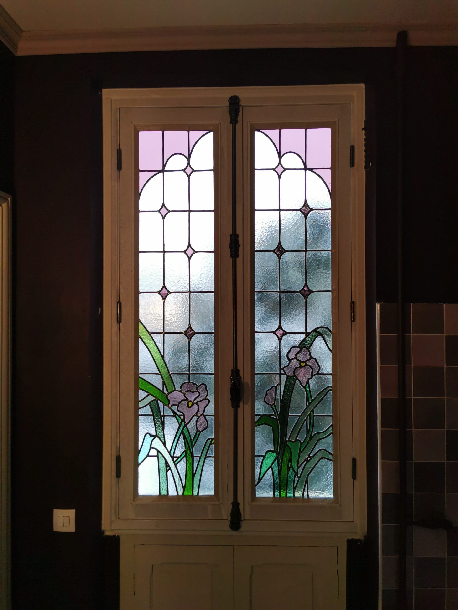 Vitrail représentant des iris violets, encadré de blanc, sur un mur brun foncé.