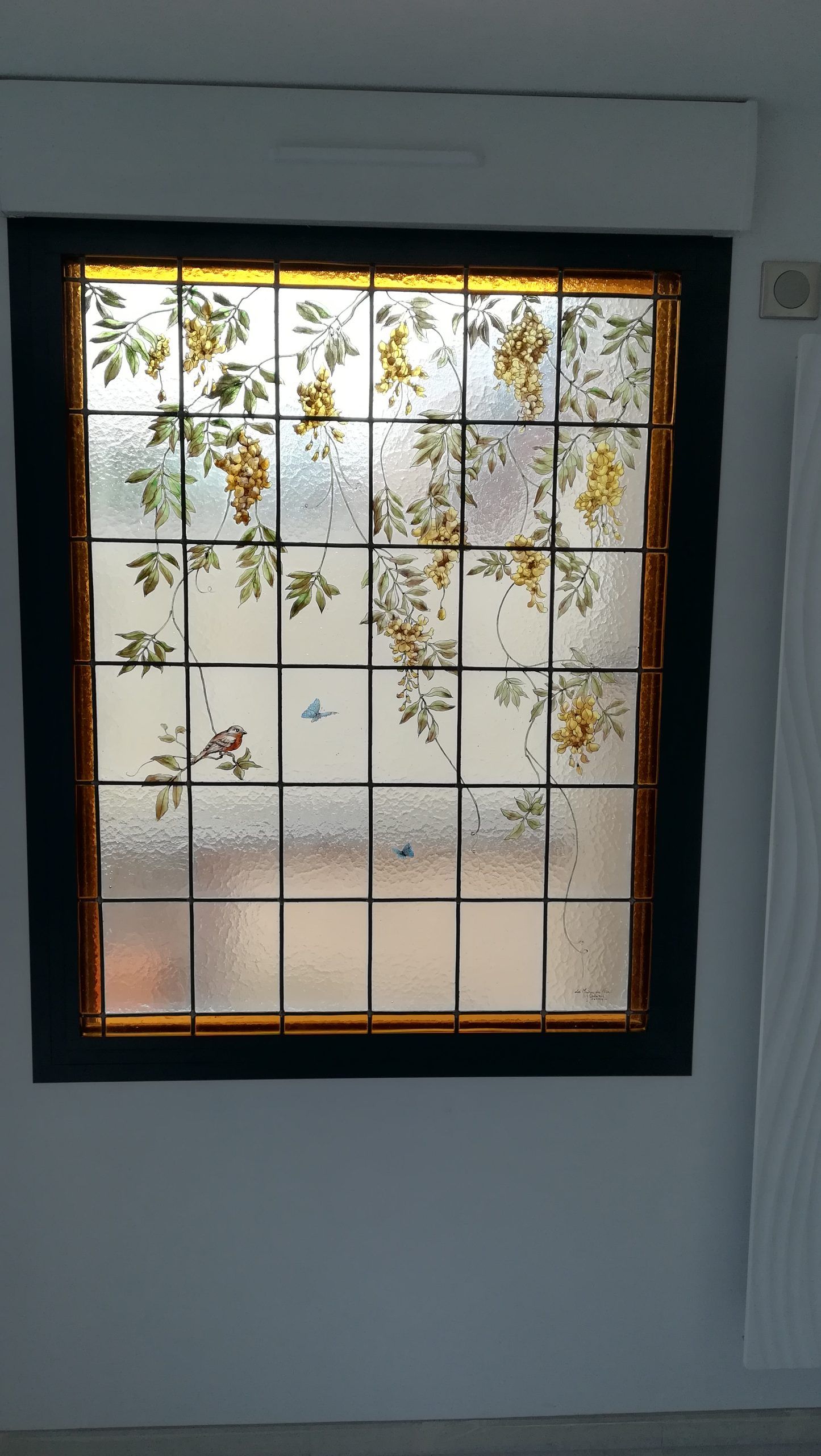 Vitrail à motif quadrillé et floral, encadré d'un cadre en bois foncé sur un mur blanc.