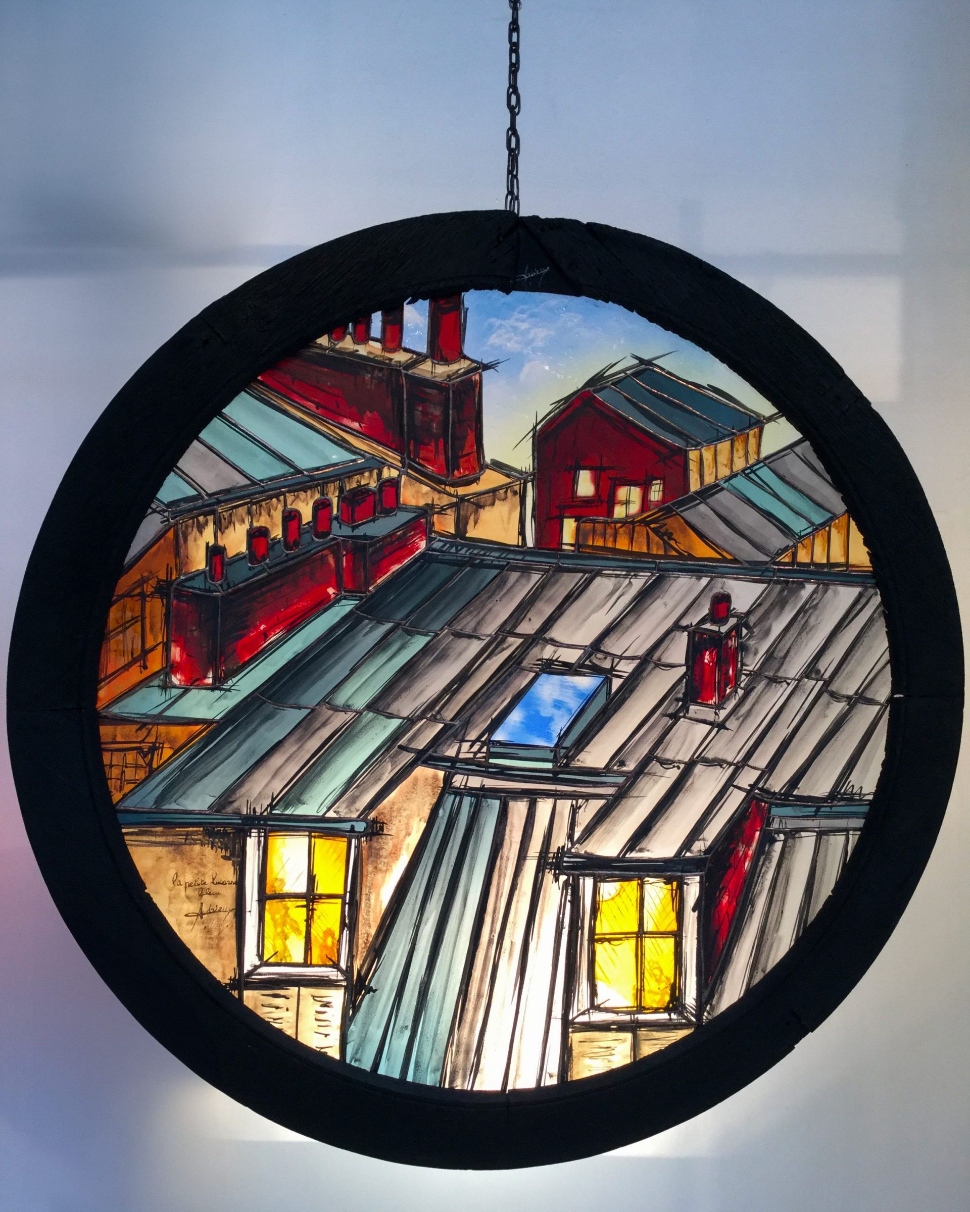 Scène de toit en vitrail dans un cadre circulaire noir, avec des cheminées rouges, un ciel bleu et des fenêtres illuminées.