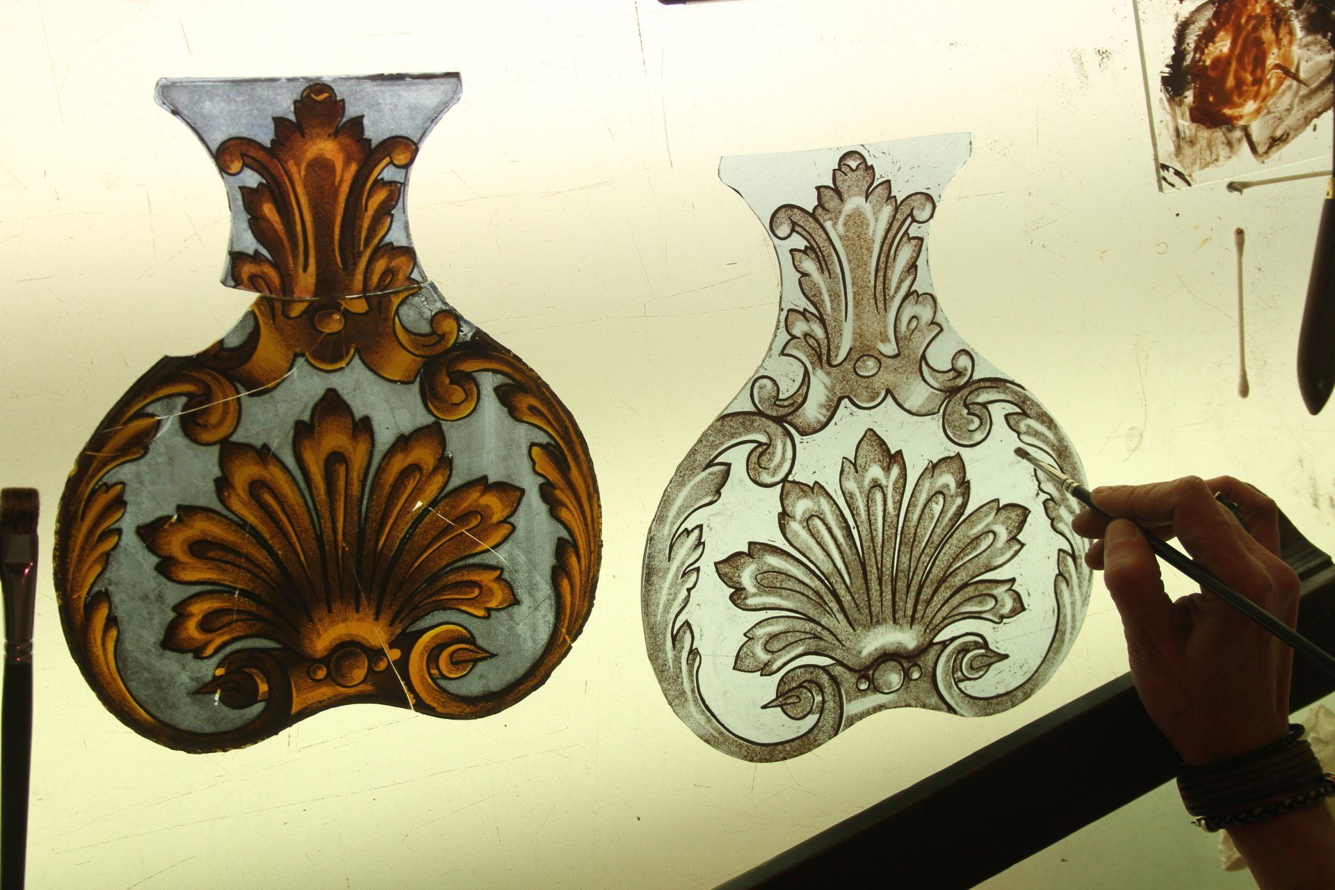 Deux motifs de vitraux, l'un coloré, l'autre aux contours dessinés. Une main peint les contours au pinceau.