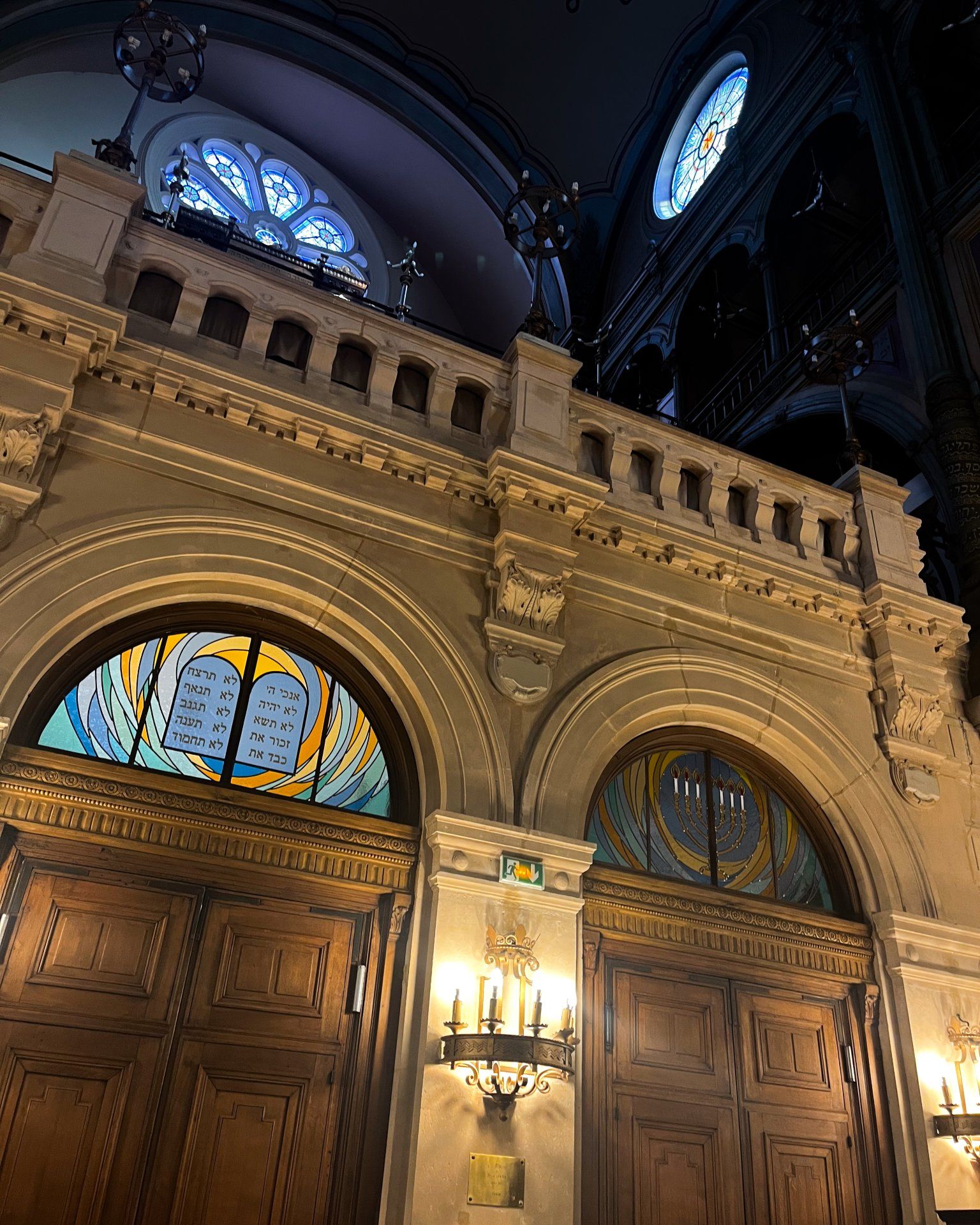 Intérieur d'une synagogue avec portes cintrées, vitraux et balcon. Éclairage tamisé.