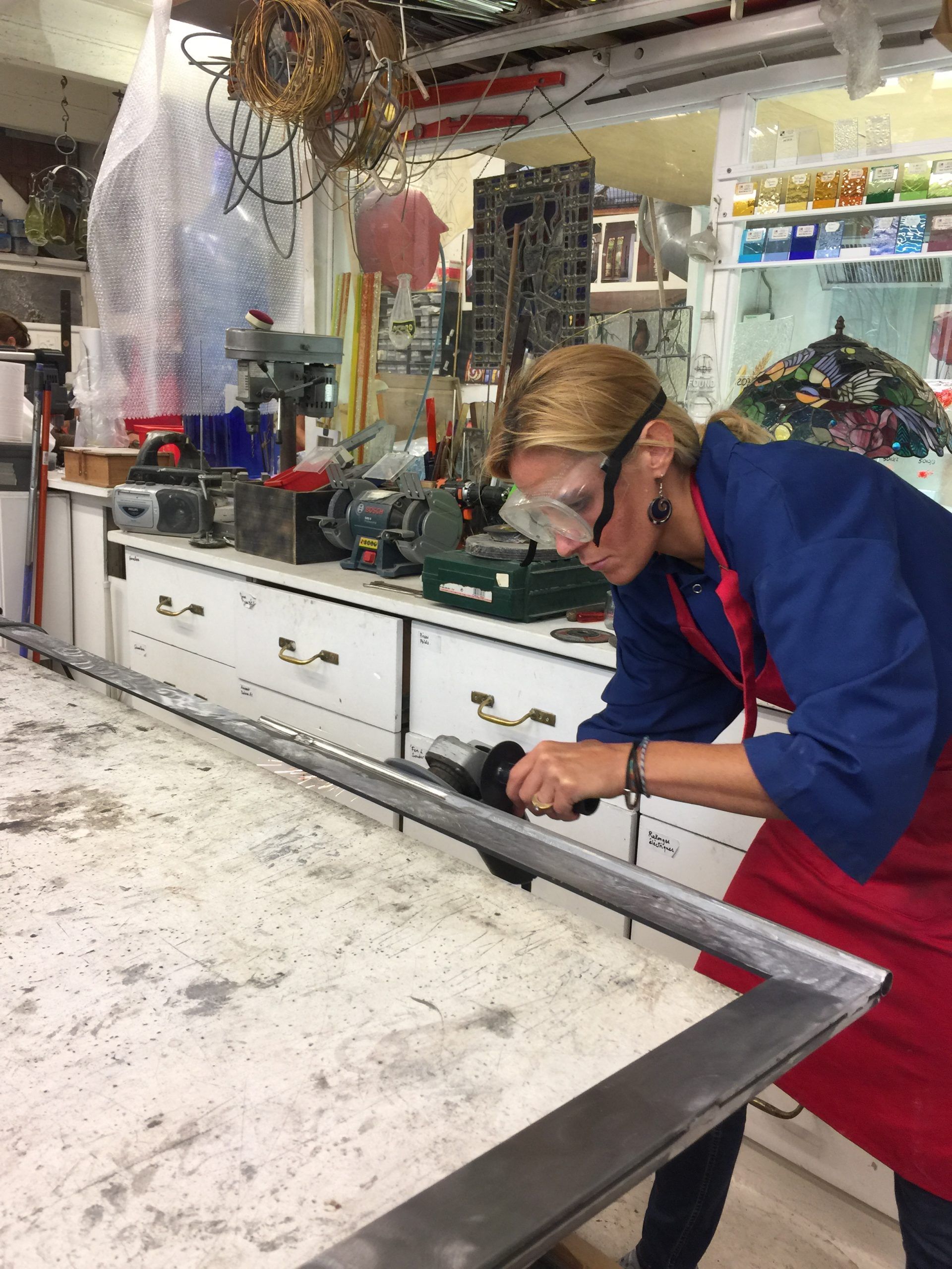Une femme portant des lunettes de sécurité utilise un outil électrique sur une structure métallique dans un atelier.